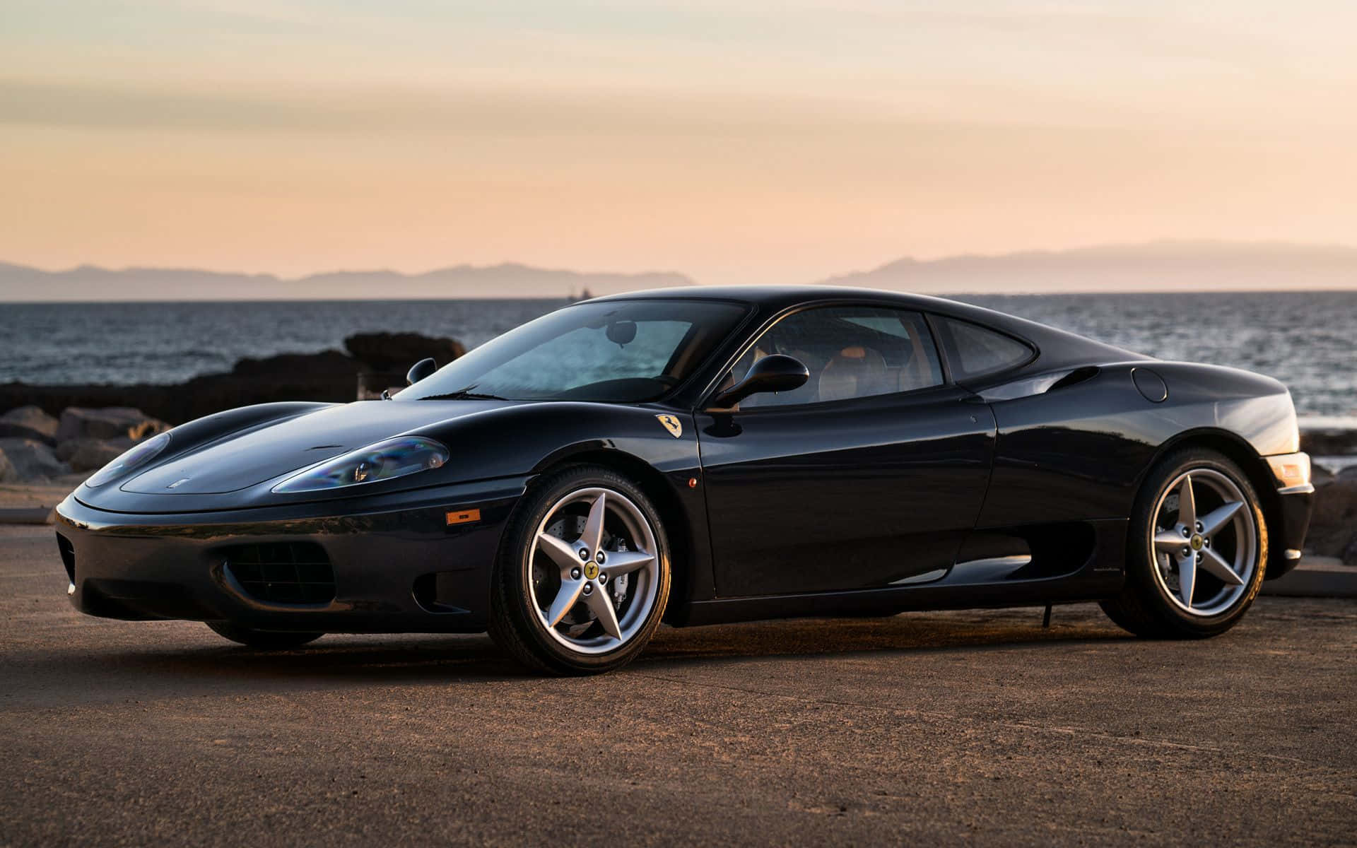 Ferrari 360 Modena 桌布 壁紙 — 一架時尚的黑色法拉利F430在日落時分停泊在沿海公路上，背景是海洋和遠處的山脈。