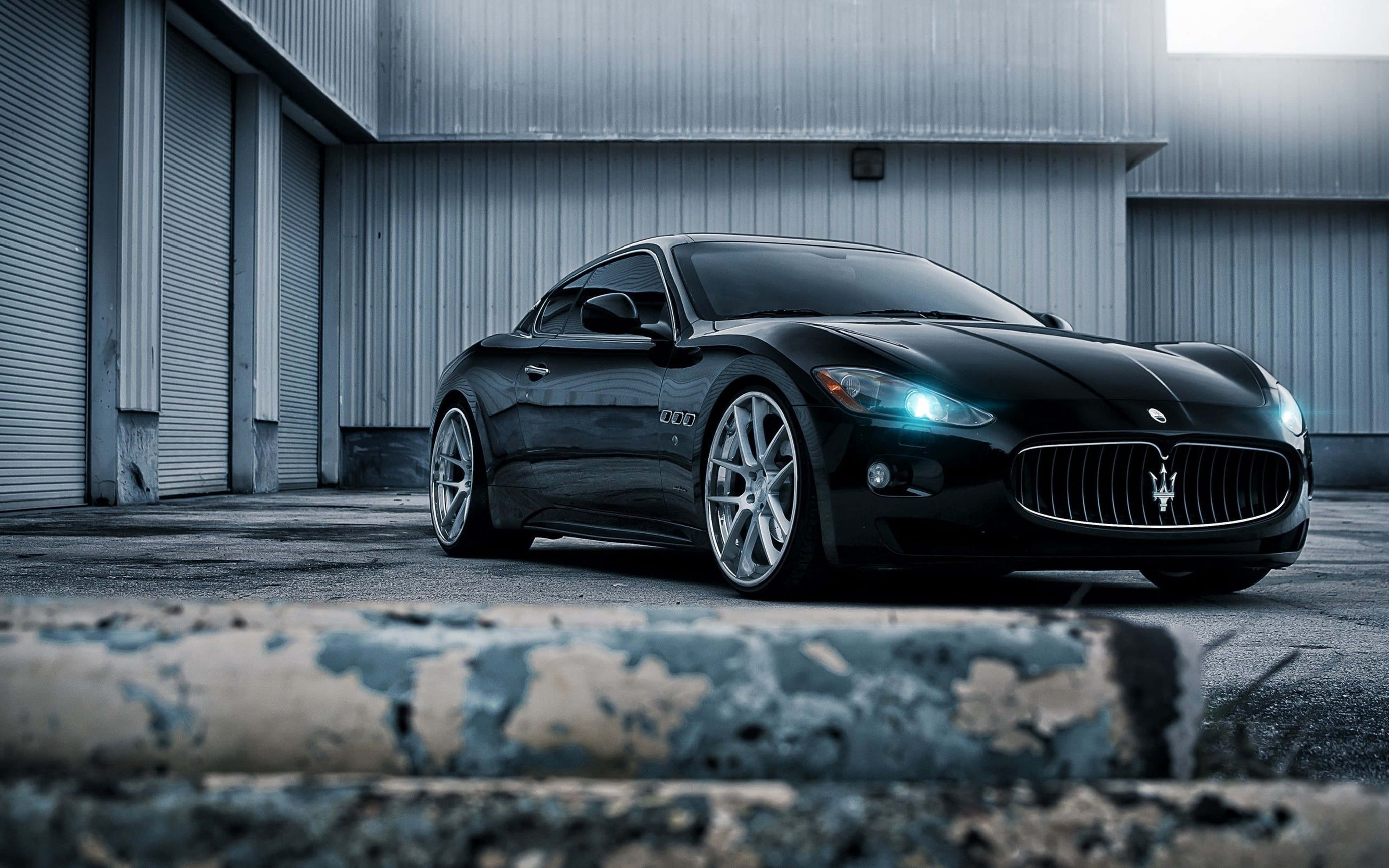 4K Maserati Low Angle 壁紙 — 一架時尚的黑色瑪莎拉蒂跑車在涼爽的燈光下閃閃發光，停在一座風化的工業建築前。