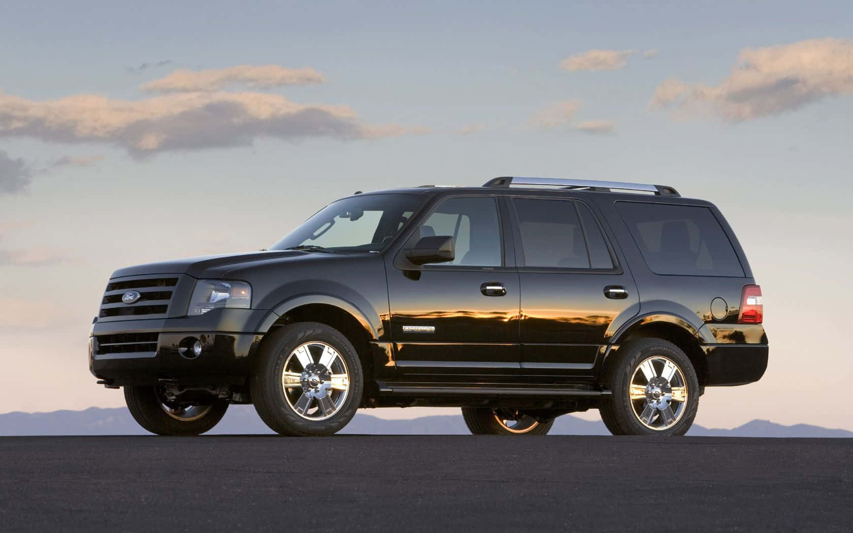 Ford Expedition 桌布 壁紙 — 一架時尚的黑色福特 Expedition SUV 停在深色瀝青路面上，背景是柔和嘅黃昏天空，遠處有山脈同散落嘅雲朵。