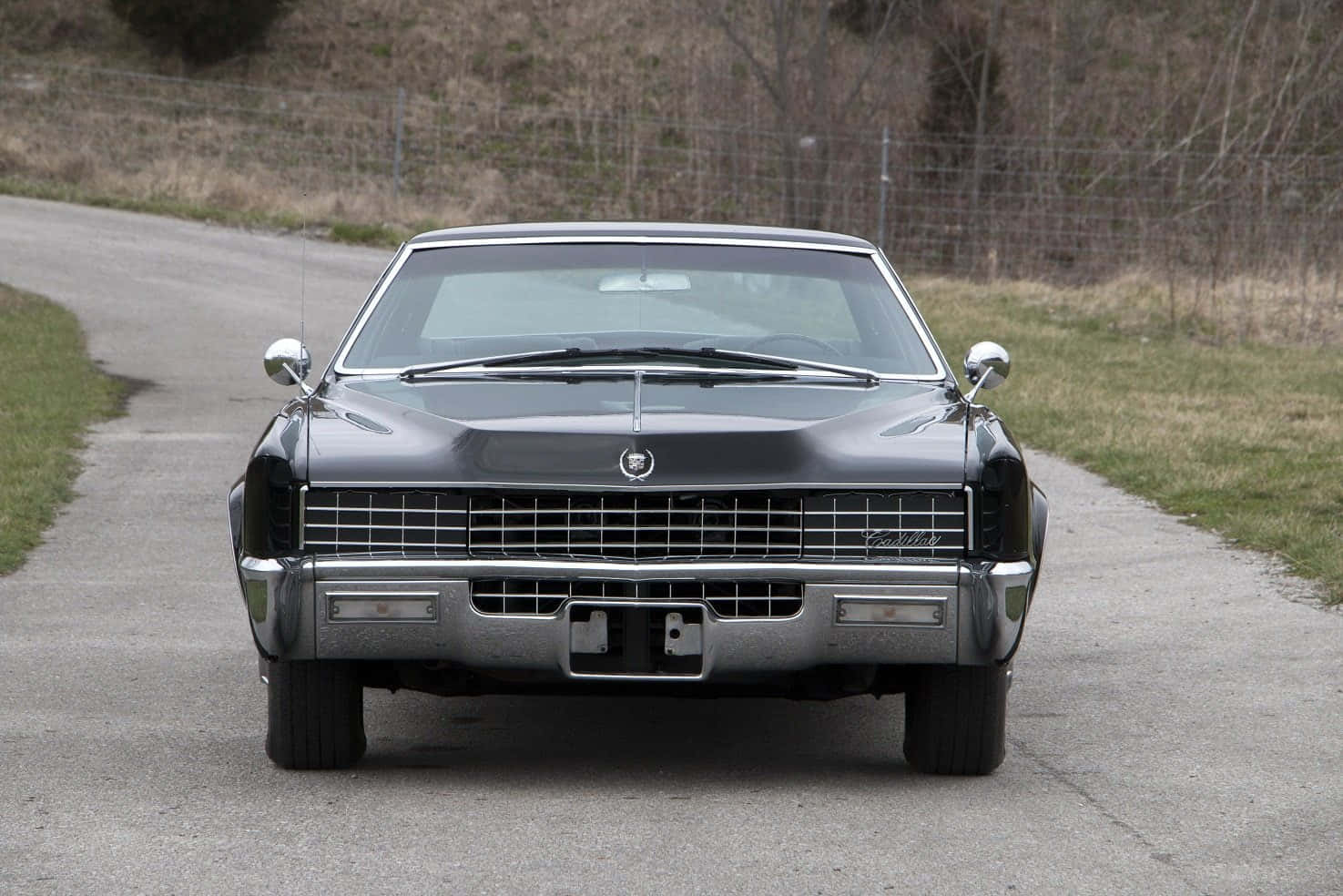 Cadillac Fleetwood 桌布 壁紙 — 一架時尚的黑色經典凱迪拉克轎車在寧靜的鄉村道路上向前行駛，兩旁是草地和光禿禿的樹木。