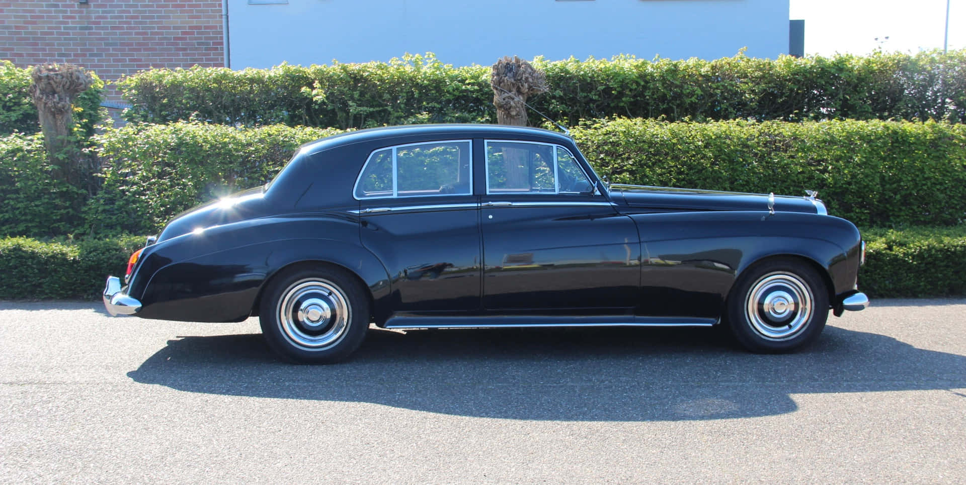 Bentley S3 桌布 壁紙 — 一架時尚的黑色經典勞斯萊斯在陽光明媚的瀝青車道上，旁邊是修剪整齊的樹籬，側面展示。