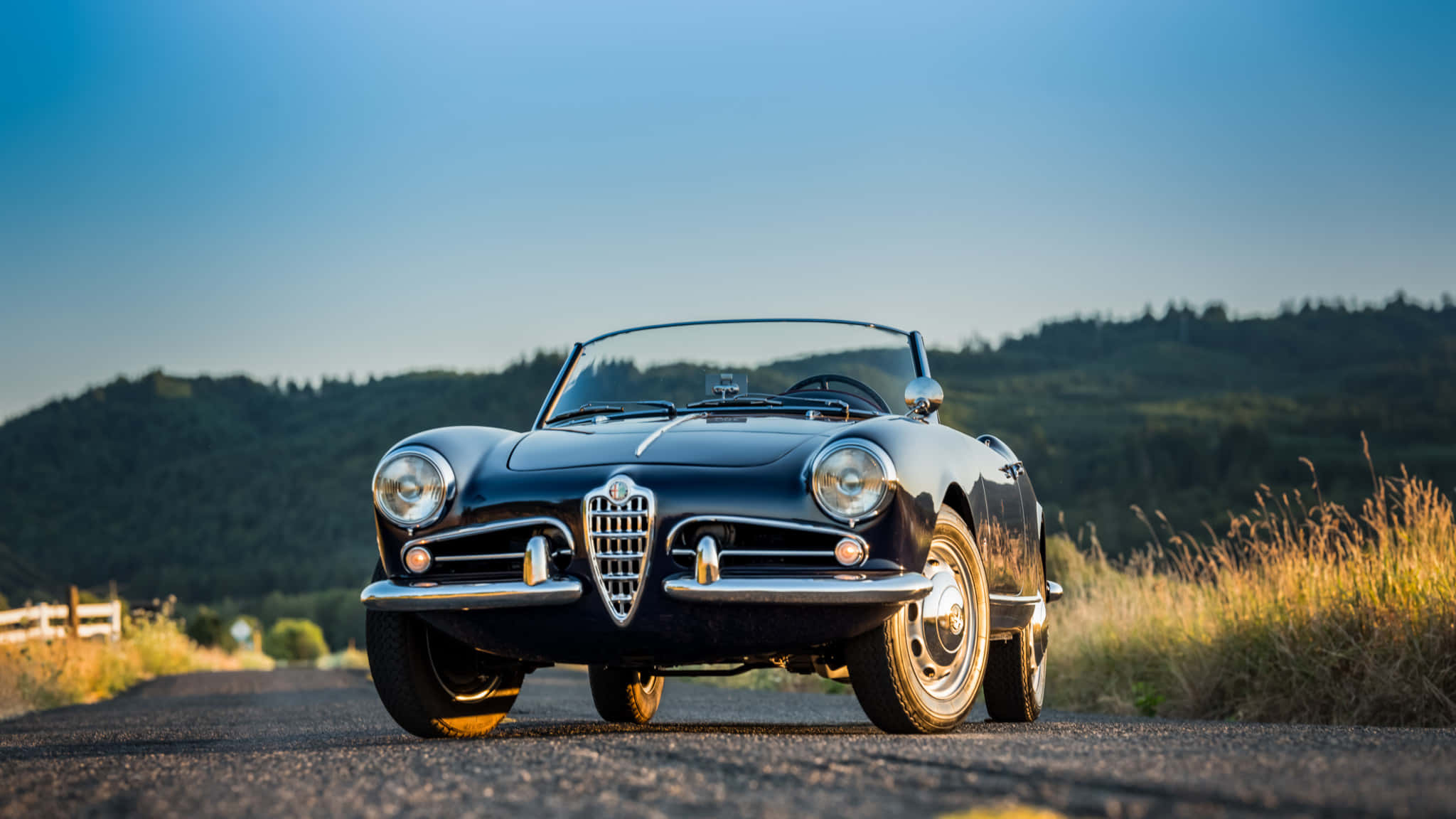 Alfa Romeo Spider 桌布 壁紙 — 一架時尚的黑色經典愛快羅密歐Spider開篷車停泊在鄉村道路上，背景是連綿起伏的綠色山丘和晴朗的藍天。
