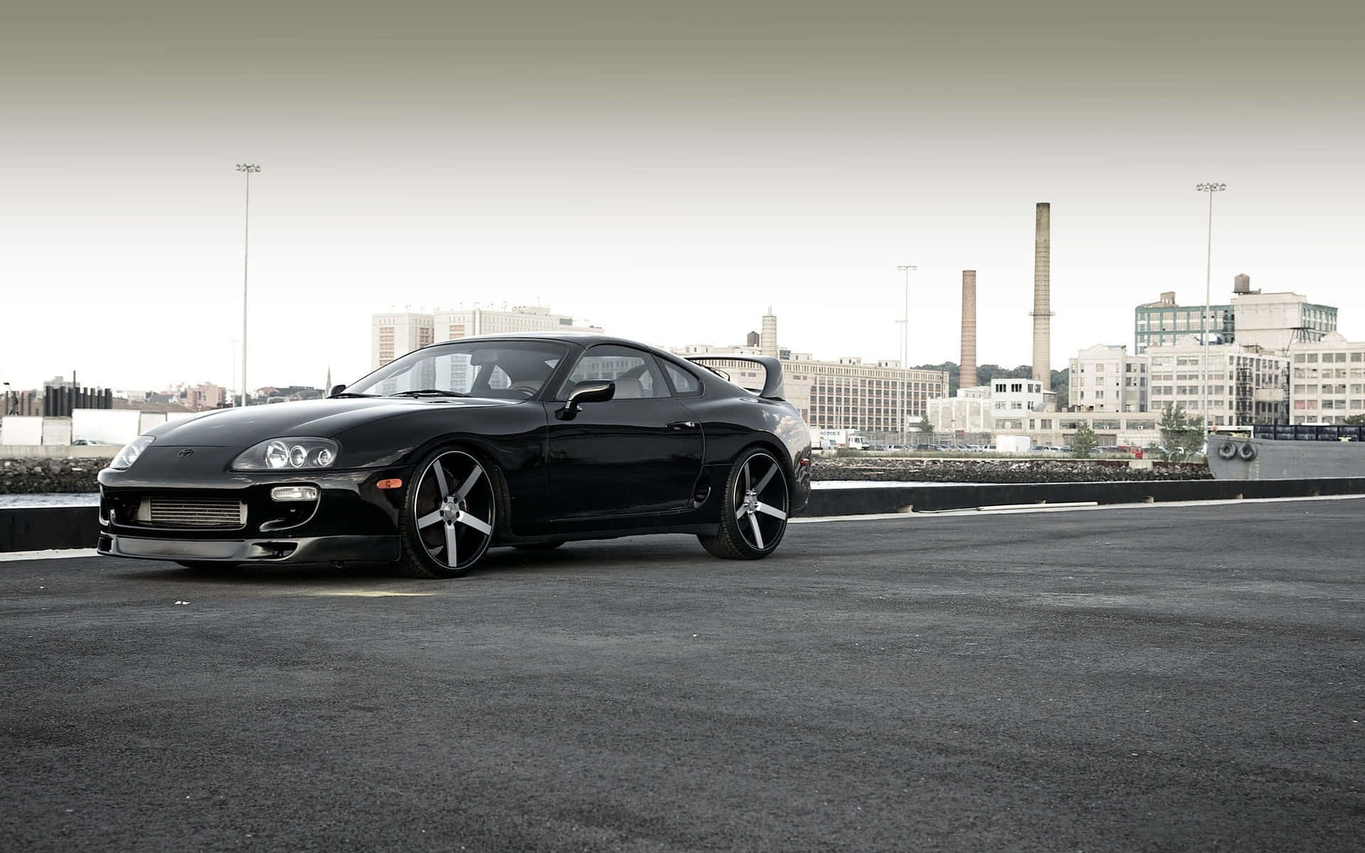 Supra 桌布 壁紙 — 一架時尚的黑色豐田Supra跑車停在瀝青停車場，背景是城市天際線和工業建築。