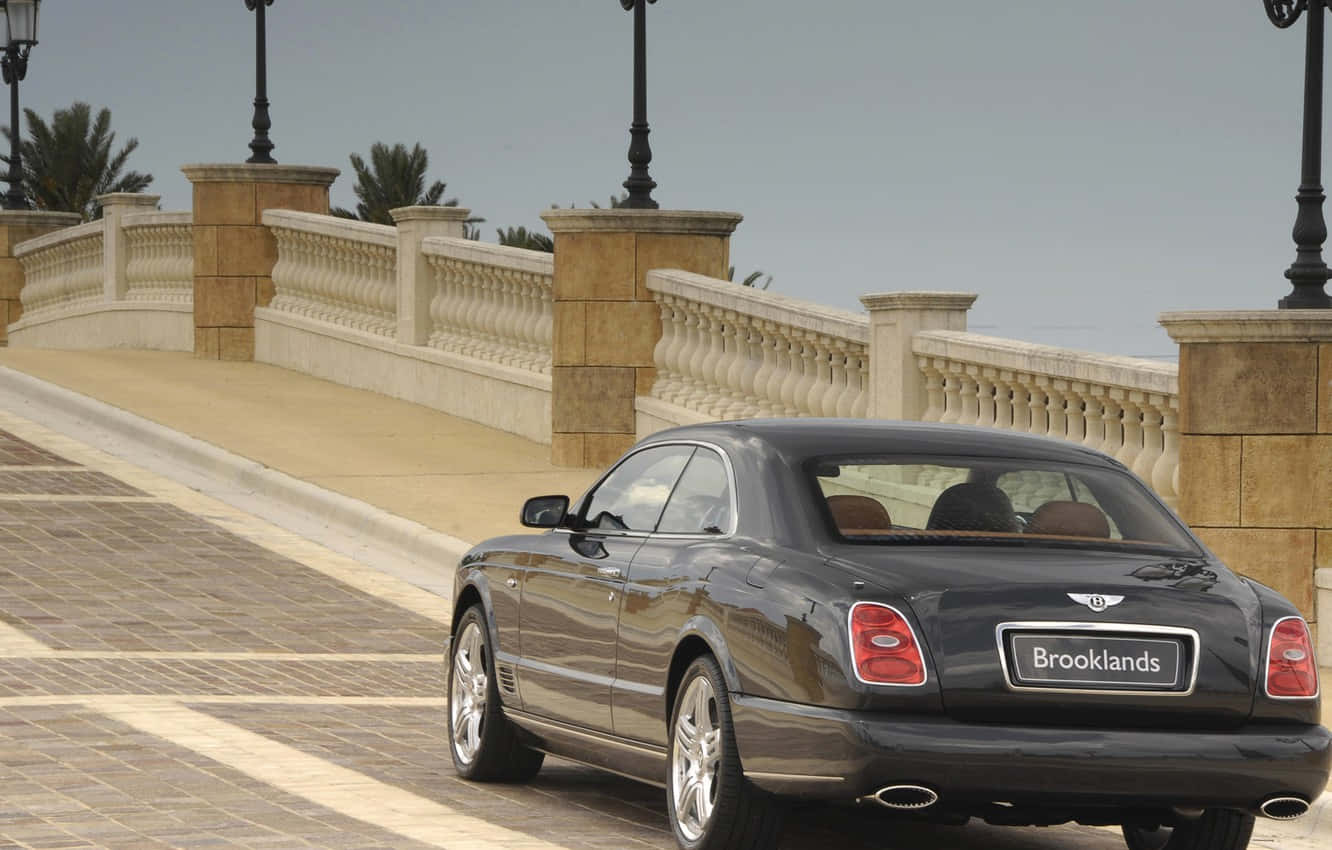 Bentley Brooklands 桌布 壁紙 — 一架時尚的黑色賓利 Brooklands 雙門轎跑車在陽光明媚的石橋上行駛，背景有華麗的欄杆和棕櫚樹。