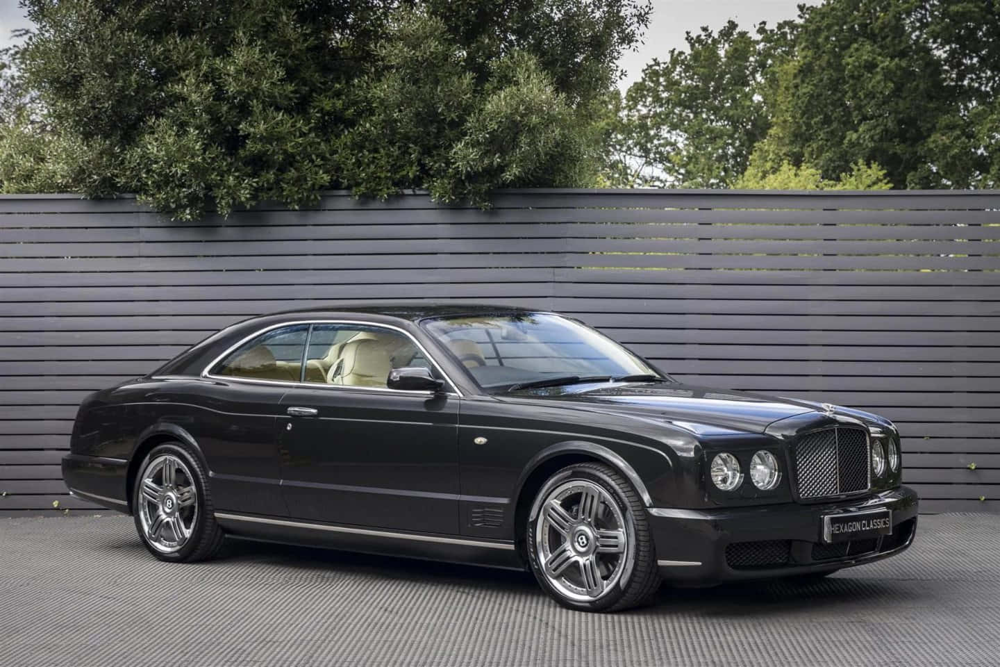 Bentley Brooklands 桌布 壁紙 — 一架時尚的黑色賓利 Continental GT 雙門跑車停在灰色網格地板上，後面有深灰色板條牆和綠色樹木。