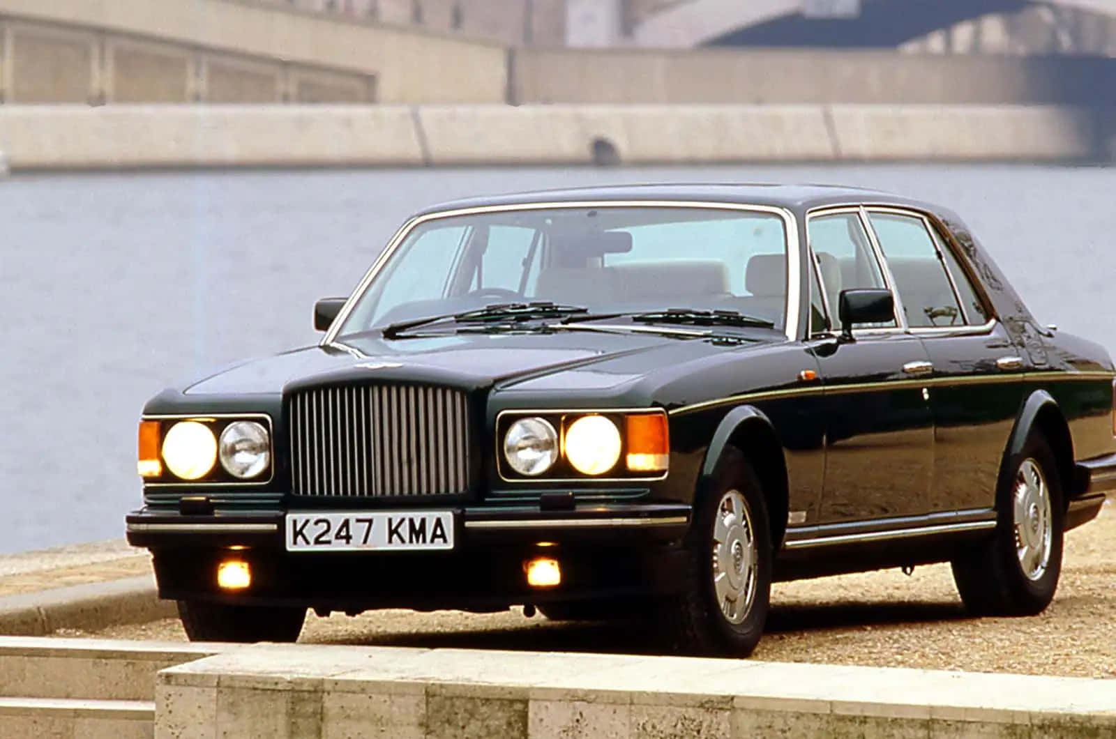 Bentley Turbo R 桌布 壁紙 — 一架時尚的黑色賓利Turbo R停在海濱，車頭燈亮起，前景有混凝土護欄。