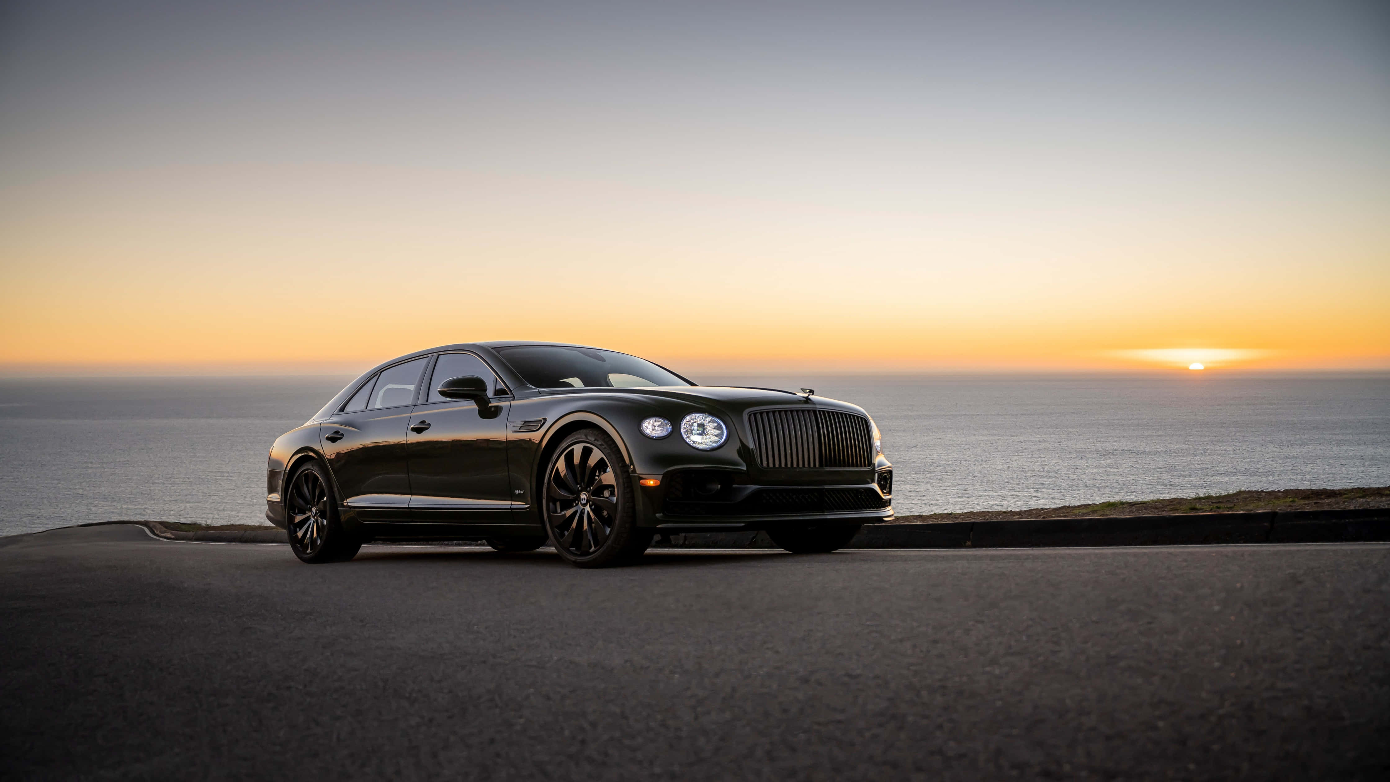 Bentley Flying Spur 桌布 壁紙 — 一架時尚的黑色賓利房車停泊在日落時分的海岸公路上，海洋地平線閃耀著橙色光芒。