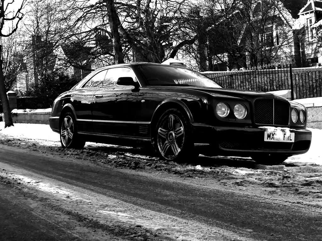 Bentley Brooklands 桌布 壁紙 — 一架時尚的黑色賓利房車停泊在積雪街道上，背景有光禿的樹木和圍欄。