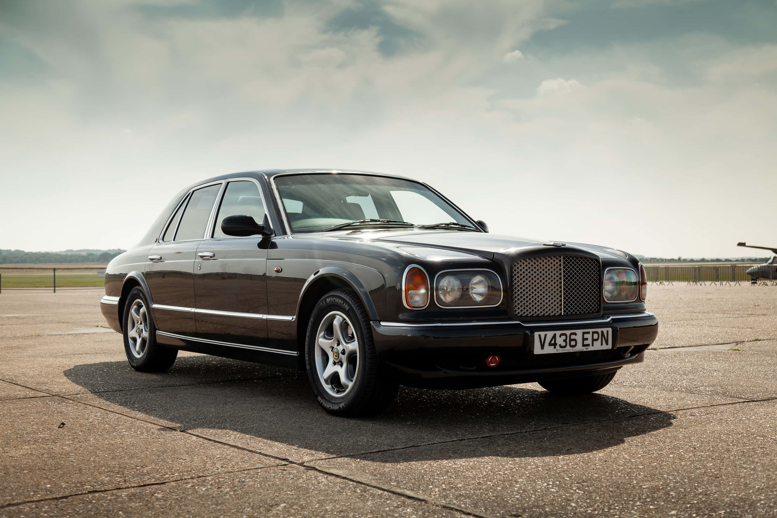 Bentley Arnage 桌布 壁紙 — 一架時尚的黑色賓利房車停泊在鋪砌的機場上，天空多雲，遠處有樹木。