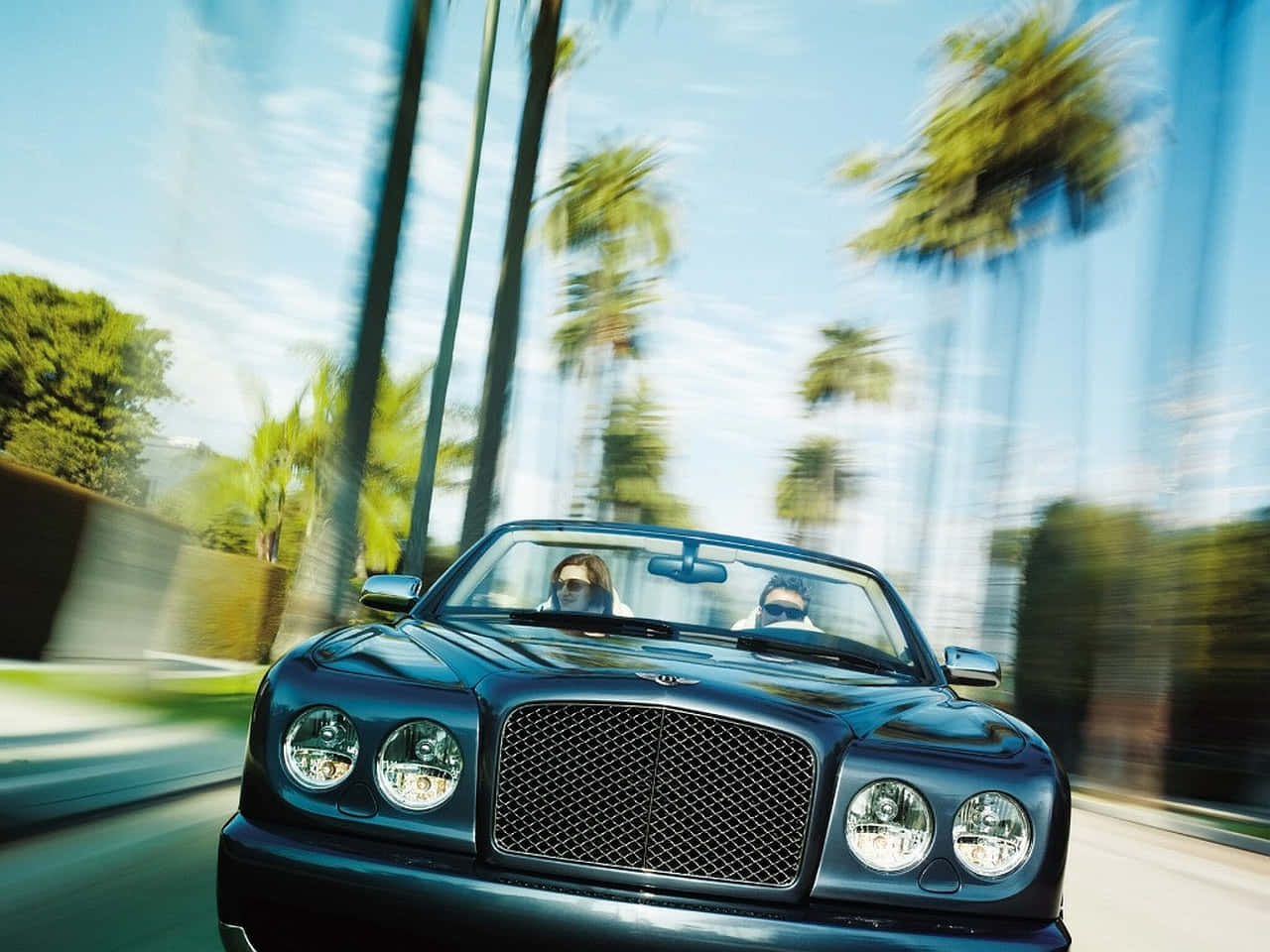 Bentley Azure 桌布 壁紙 — 一架時尚的黑色賓利開篷跑車在棕櫚樹成蔭的道路上行駛，車上兩人戴著太陽眼鏡，享受動態模糊的效果。