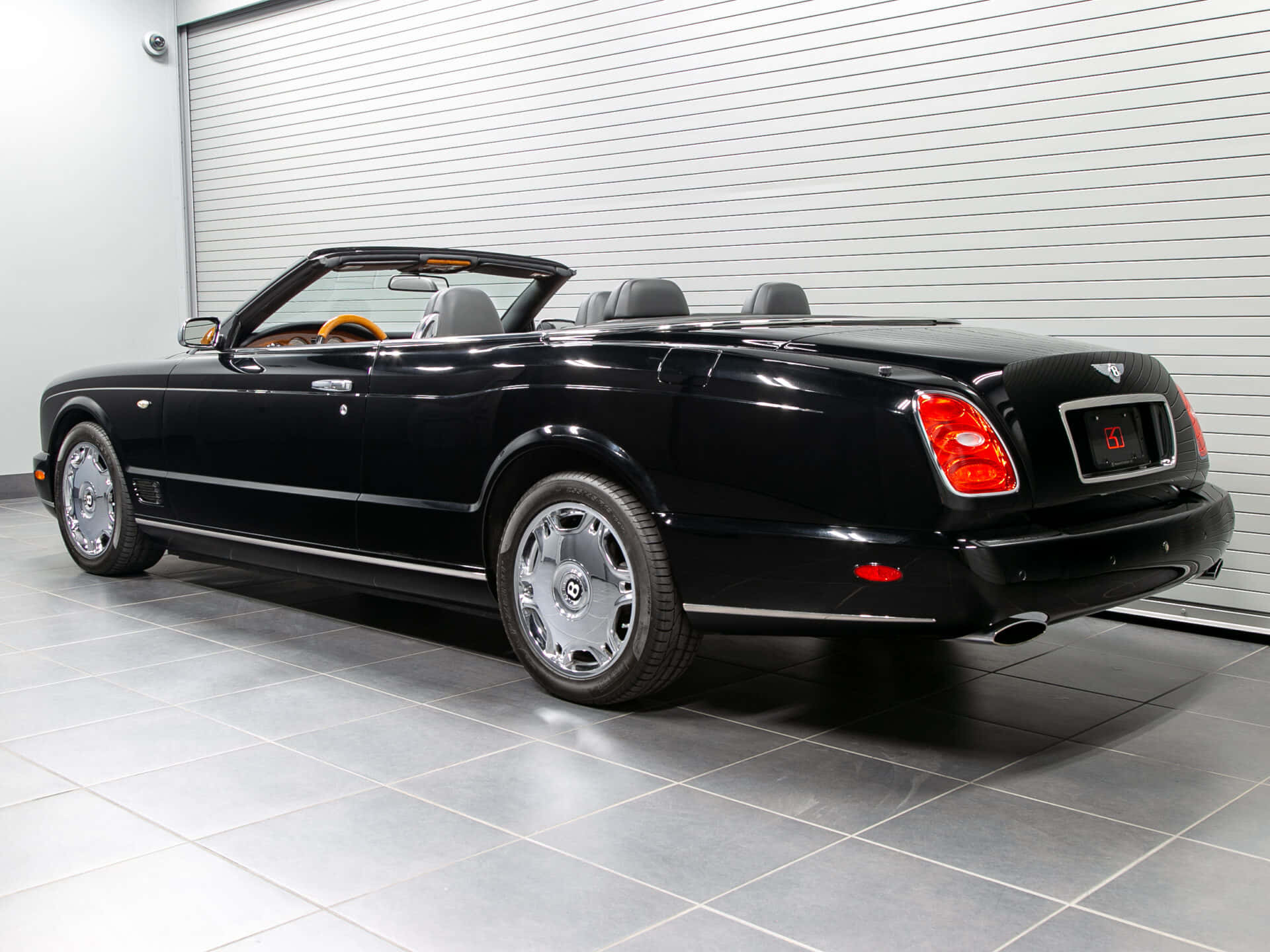 Bentley Azure 桌布 壁紙 — 一架時尚的黑色賓利開篷車，配備鉻合金輪圈和橙色內飾，停放在一個乾淨的瓷磚車庫中，車庫有白色捲簾門。