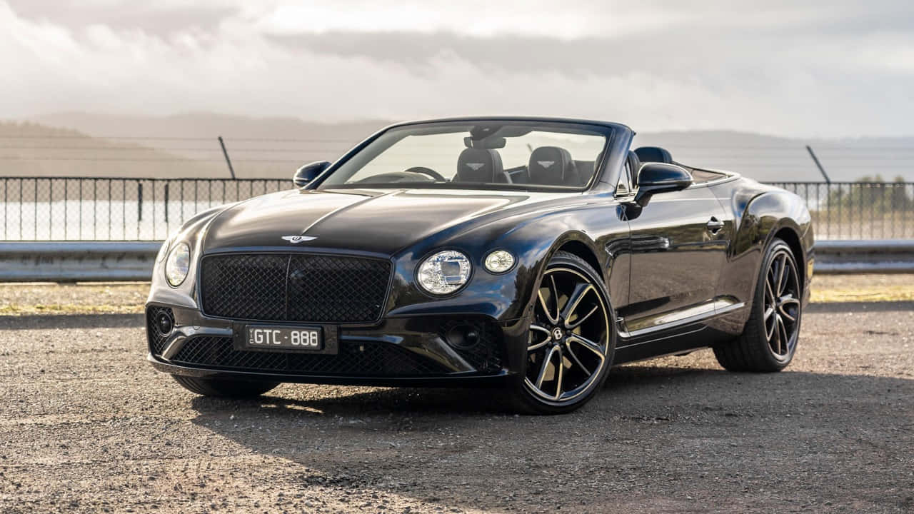 Bentley Continental Gt Convertible 桌布 壁紙 — 一架時尚的黑色賓利開篷車停泊在碎石路上，旁邊有圍欄觀景點和遠處的山丘，天空多雲。
