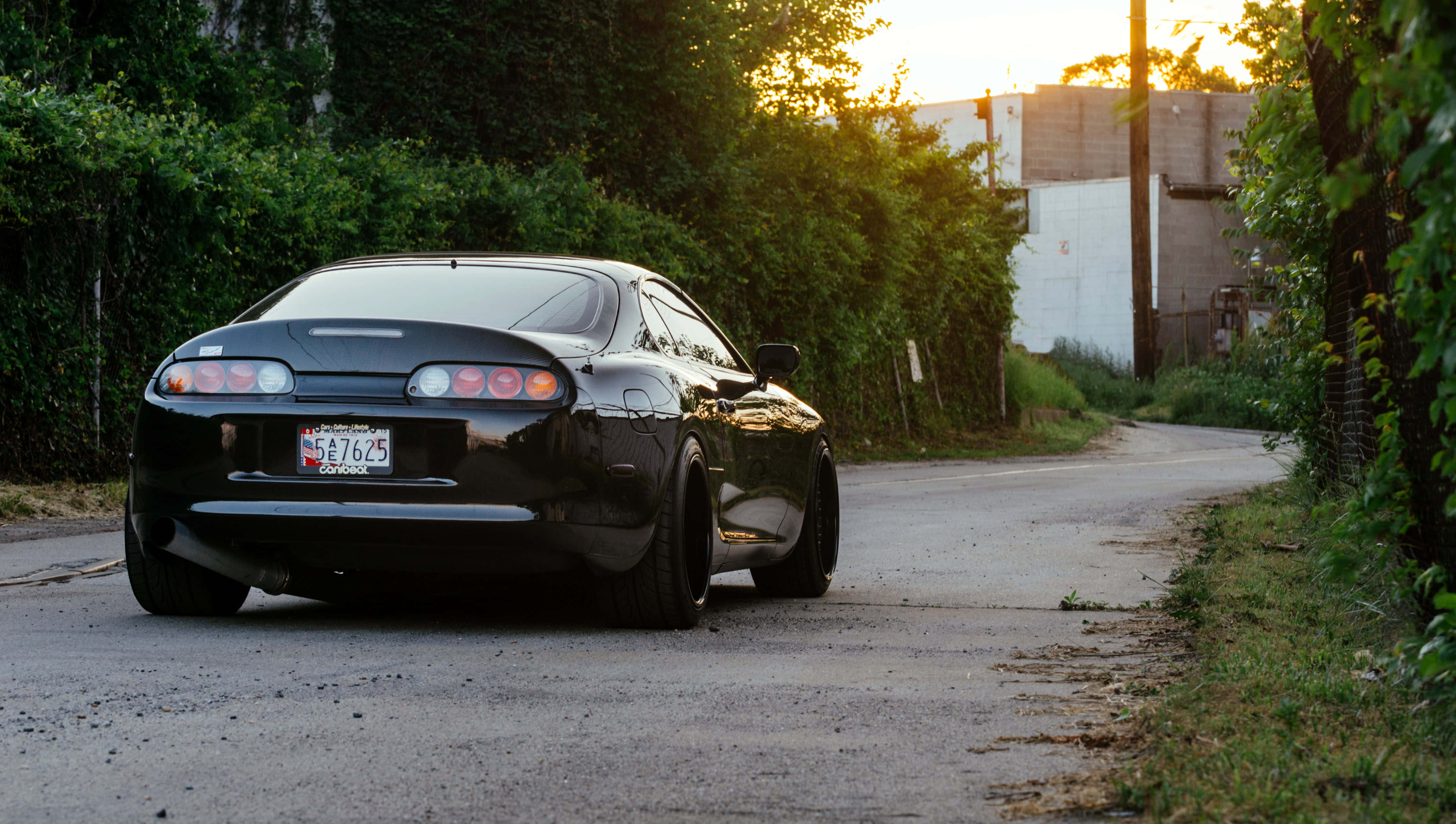 Supra 桌布 壁紙 — 一架時尚的黑色跑車停在寧靜的瀝青路上，正值黃金時刻，周圍環繞著茂密的綠植和柔和的光線。