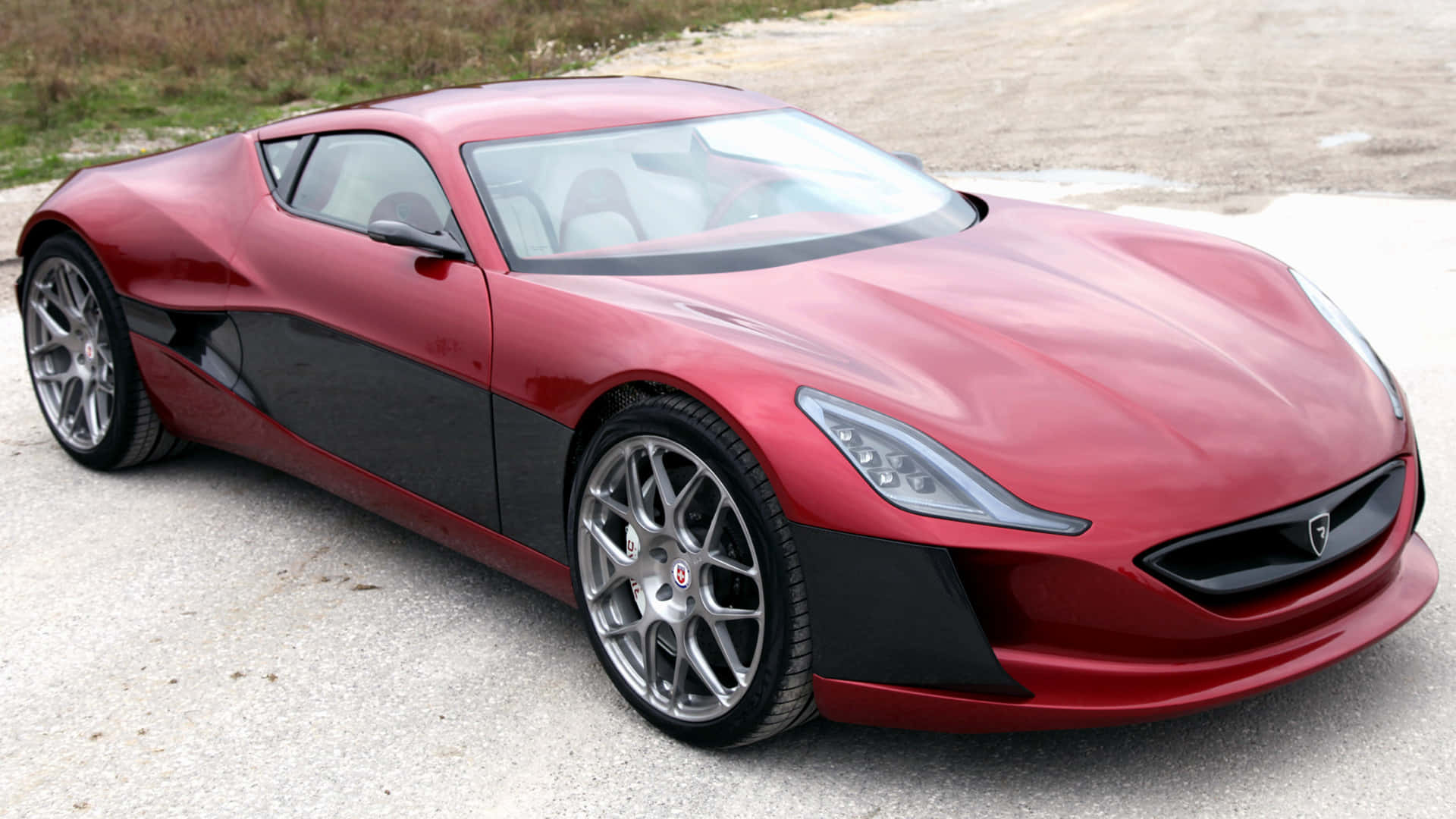 Rimac 桌布 壁紙 — 一架時尚紅黑配色、銀色輪圈的跑車，在明亮日光下停泊在碎石路面上。