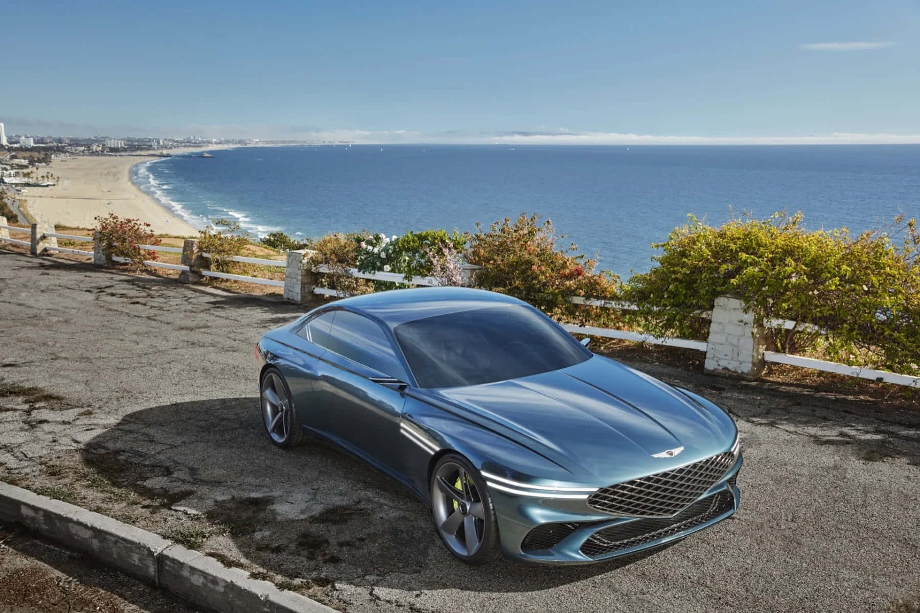 Genesis X Concept 桌布 壁紙 — 一架時尚金屬藍色跑車停泊在海岸懸崖上，俯瞰著沙灘和廣闊的海洋，天空清澈湛藍。