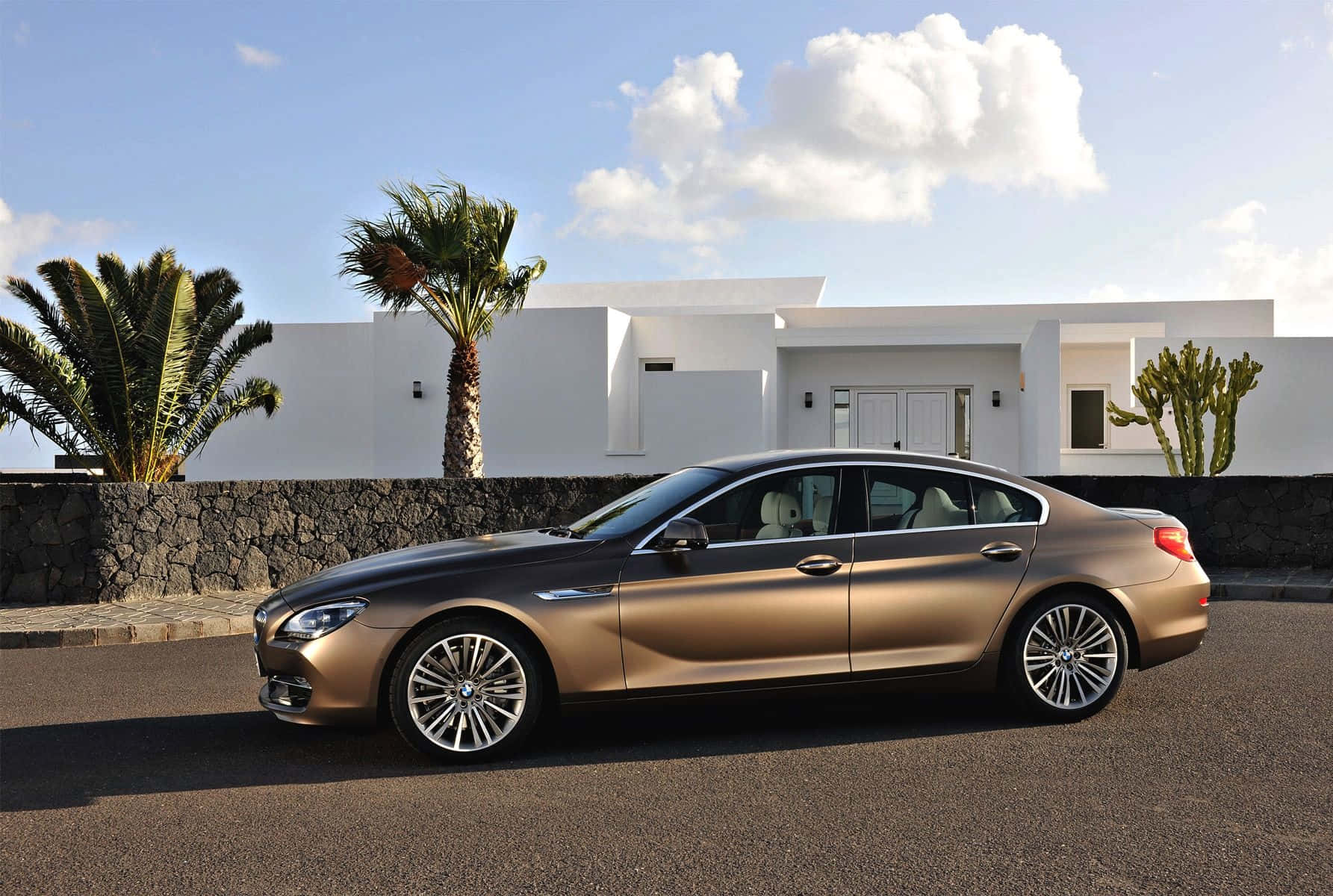 Bmw 6 Series 桌布 壁紙 — 一架時尚金色寶馬房車停泊在現代白色別墅前的瀝青路上，別墅周圍有棕櫚樹，上方是明亮的藍天。