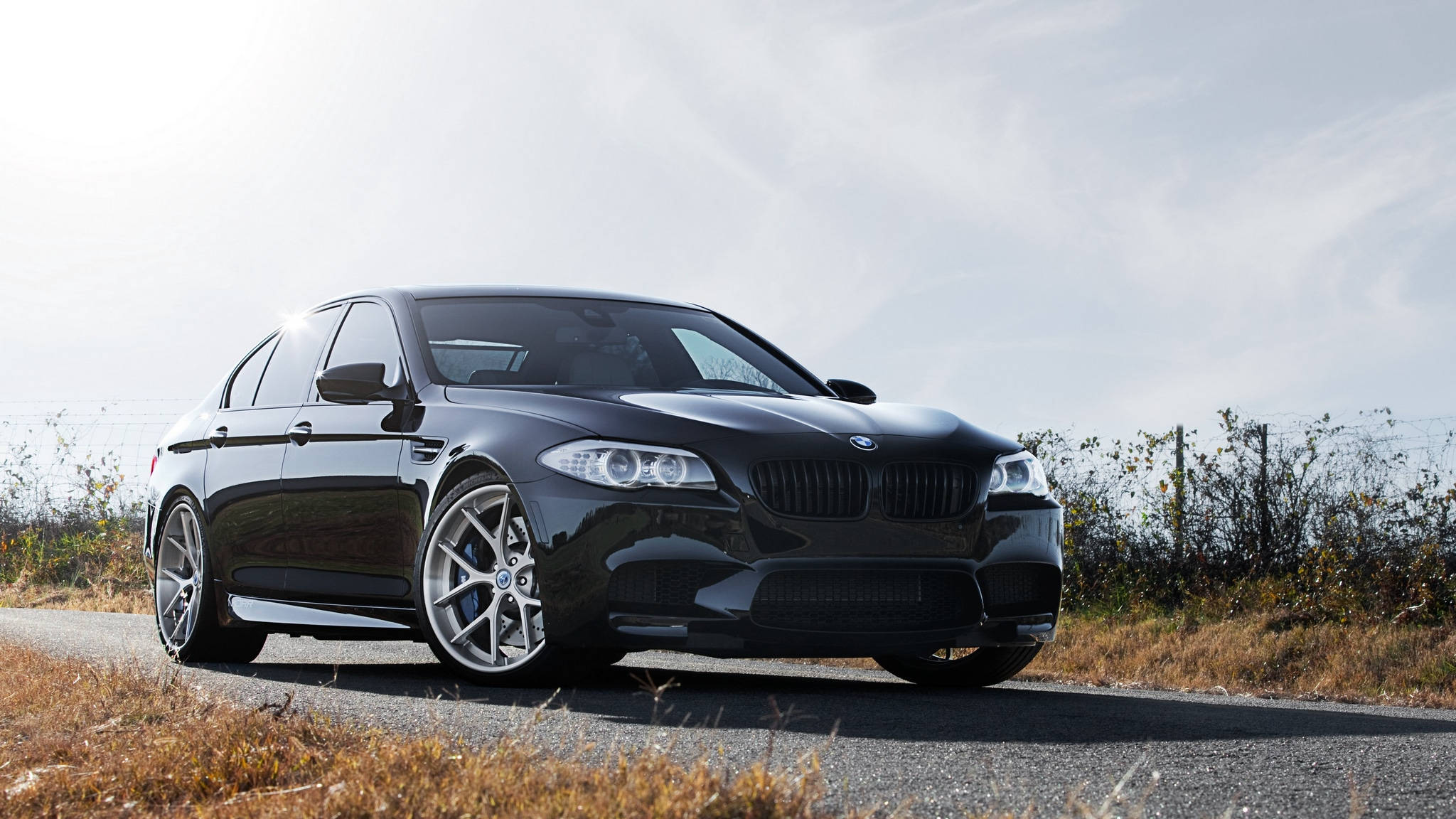 黑色動態圖 桌布 壁紙 — 一架時尚黑色BMW M5運動轎車停泊在乾草和陰天嘅鄉村道路上。