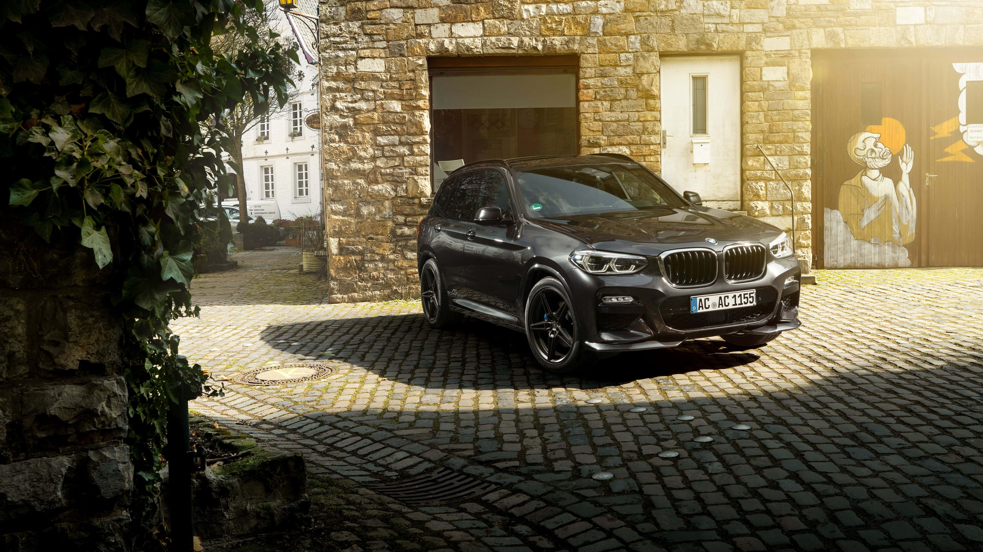 Bmw X3 桌布 壁紙 — 一架時尚黑色BMW X3 SUV停泊在陽光普照的鵝卵石庭院，旁邊是帶有常春藤和塗鴉藝術的石建築。