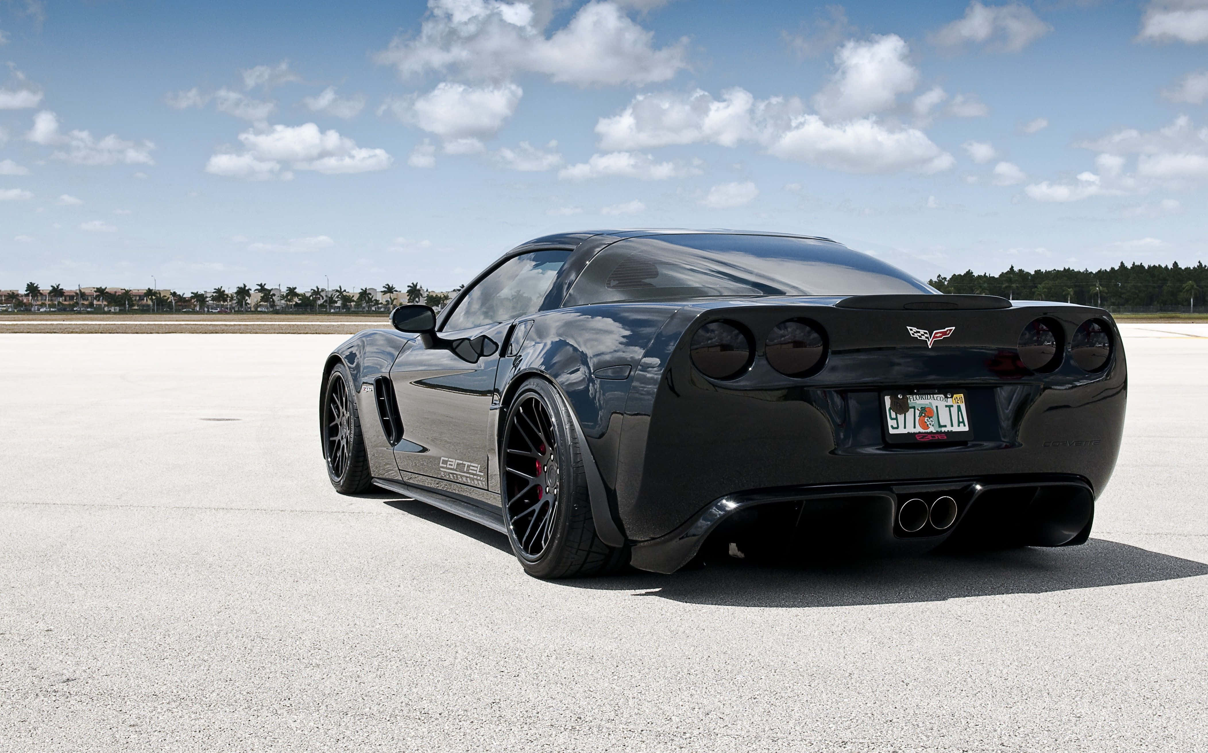 Chevrolet Corvette Z06 桌布 壁紙 — 一架時尚黑色Corvette跑車停泊在廣闊混凝土場地上，背景是晴朗藍天與零星白雲。