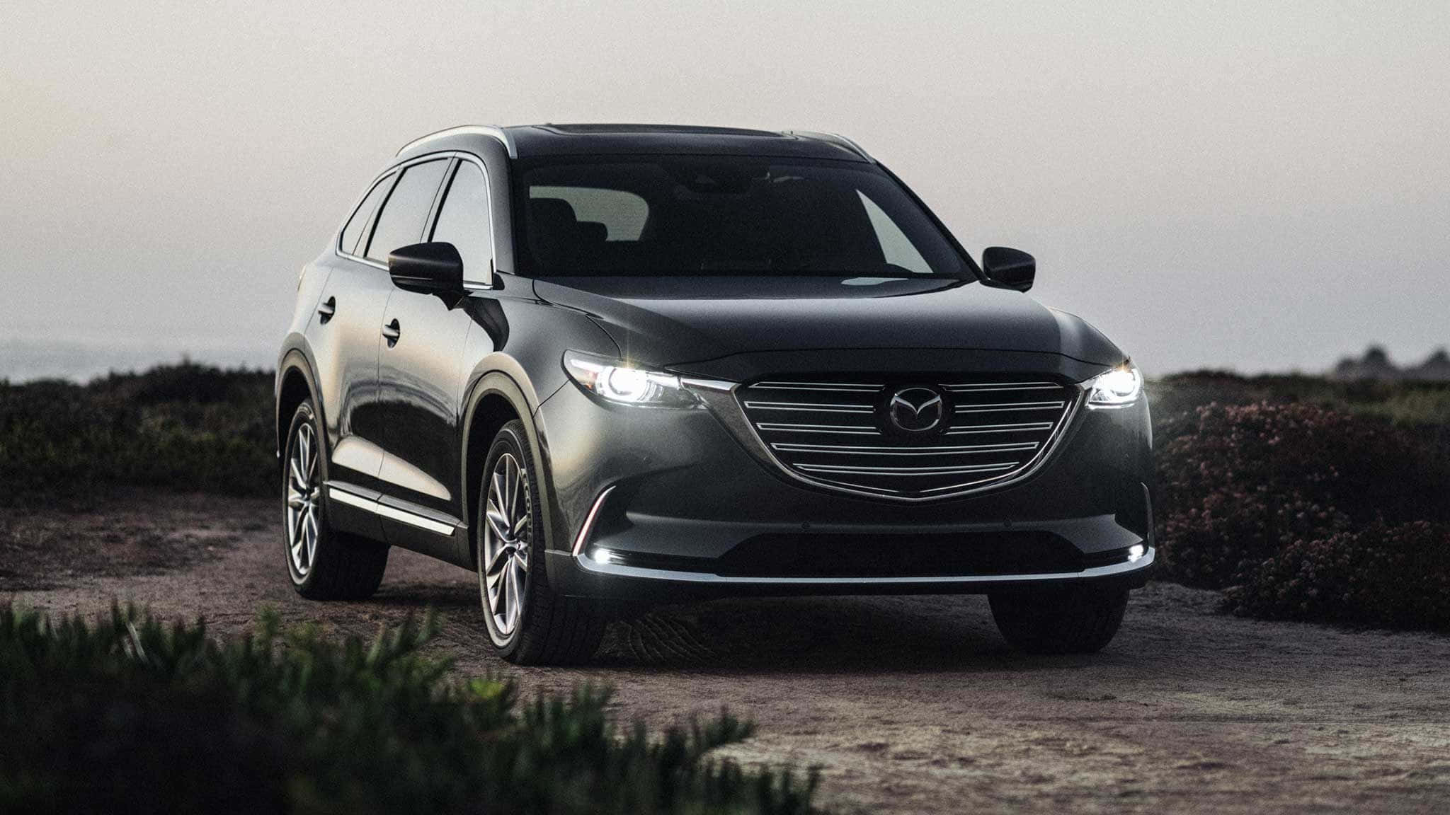 Mazda Cx-9 桌布 壁紙 — 一架時尚黑色Mazda CX-9 SUV，亮著車頭燈，停泊在塵土小徑旁的低矮灌木叢邊，天空朦朧。