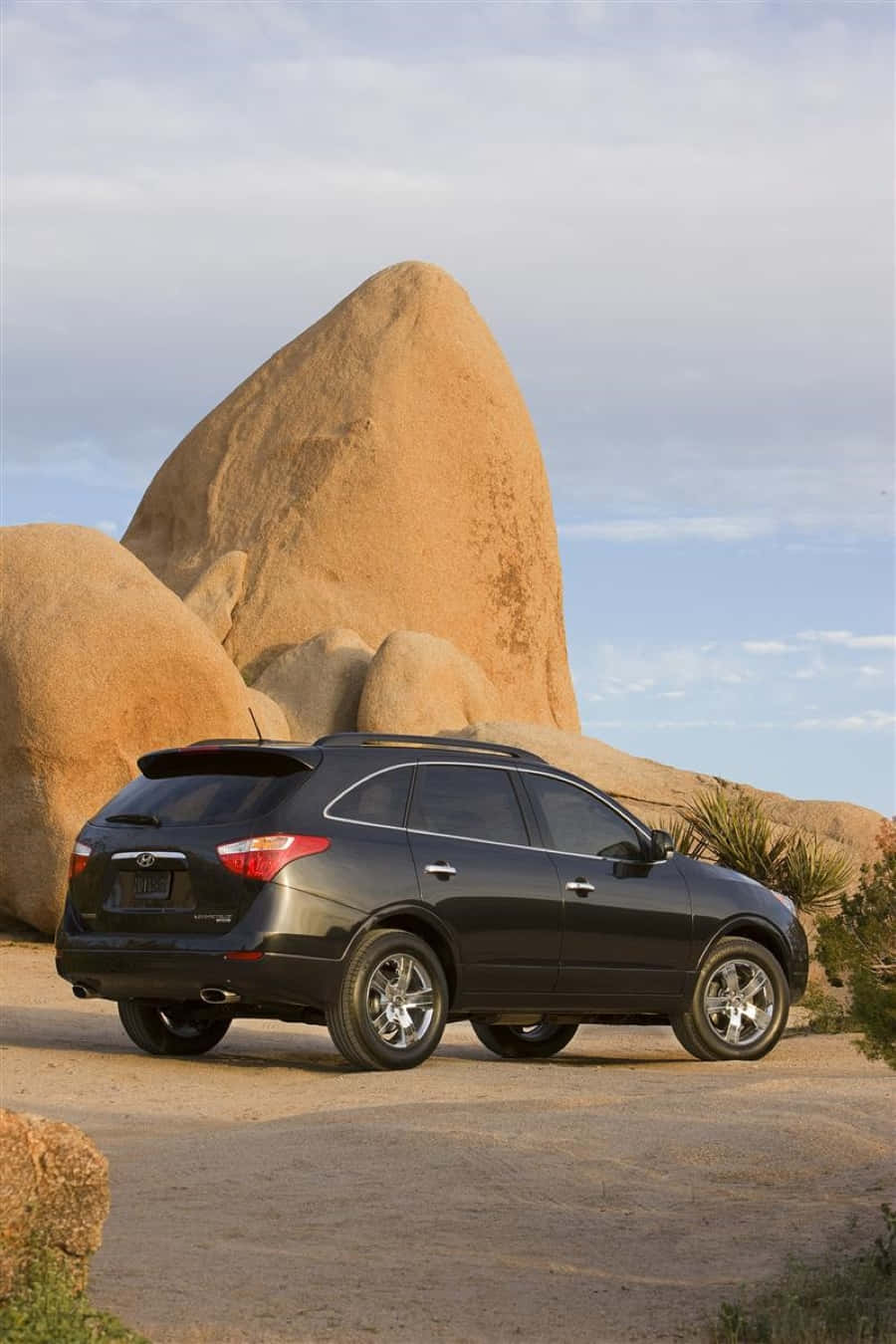 Hyundai Veracruz 桌布 壁紙 — 一架時尚黑色SUV停泊在塵土飛揚的小徑上，旁邊是巨大的棕褐色巨石，柔和藍天中散佈著雲朵。