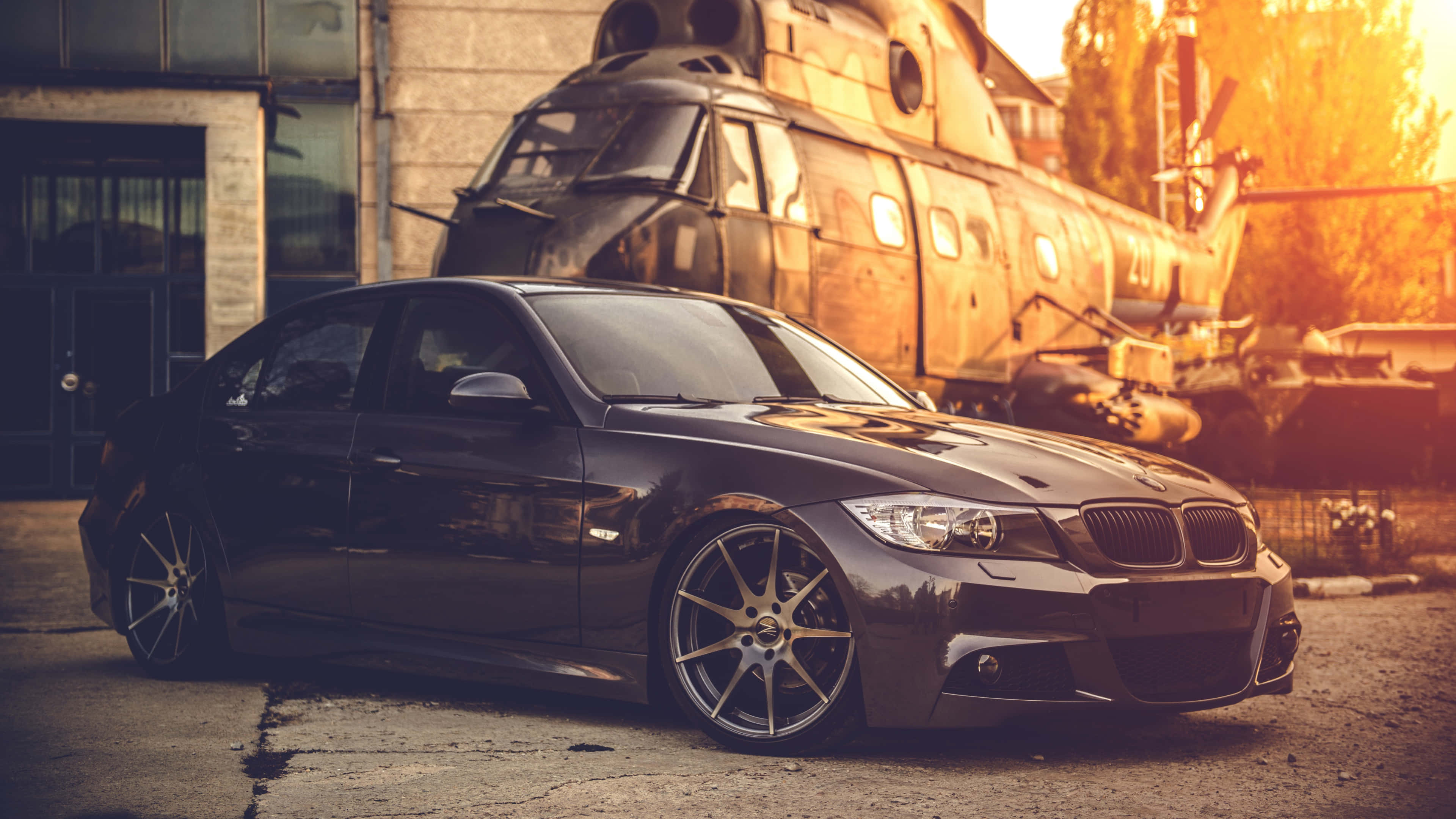 4K BMW 壁紙 桌布 壁紙 — 一架時尚黑色寶馬房車配備定制輪圈，在黃金時段停泊在一架生鏽的軍用直升機前。