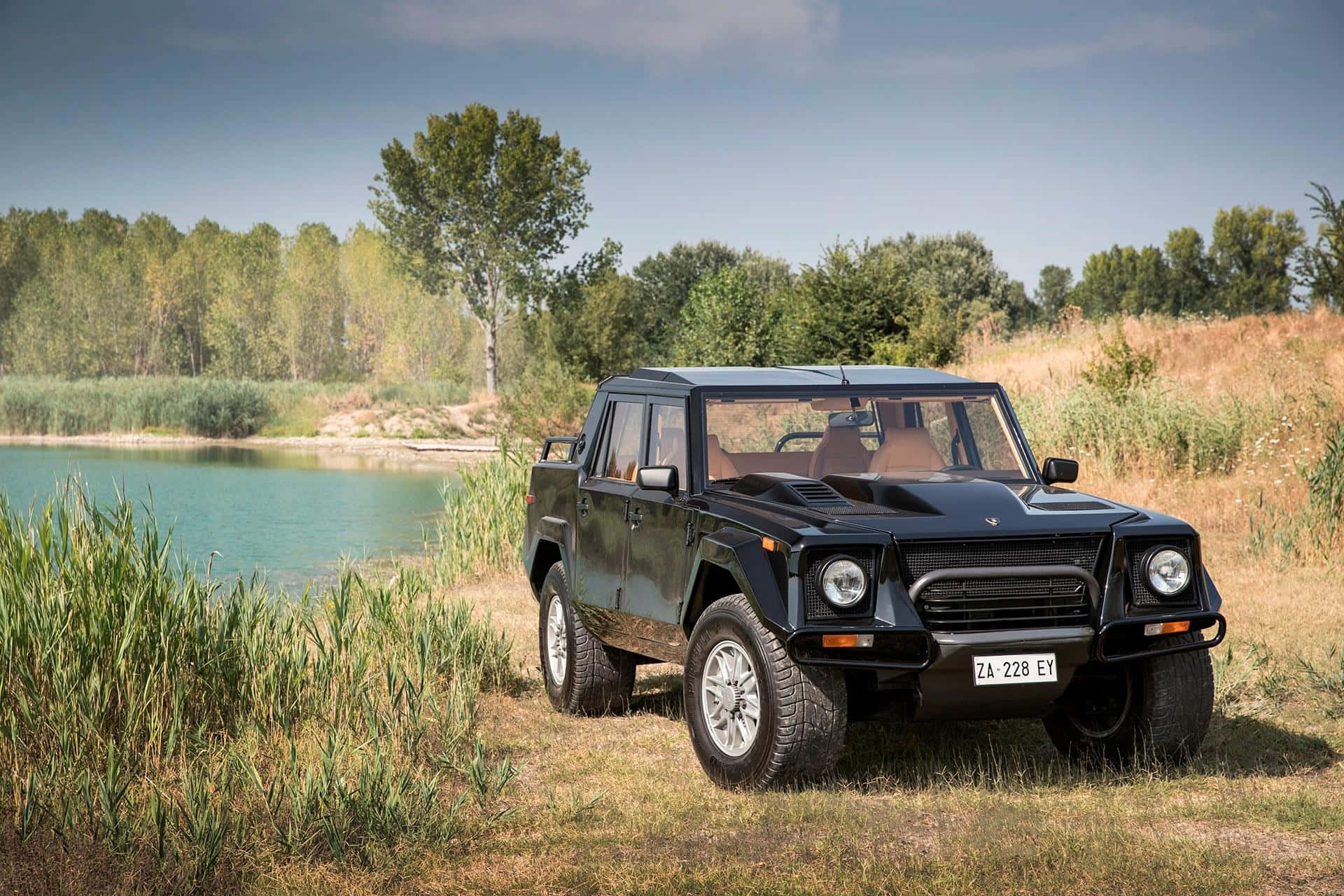 Lamborghini Lm002 桌布 壁紙 — 一架時尚黑色林寶堅尼LM002 SUV停泊在寧靜的湖邊，四周被高高的草叢和樹木環繞，天空清澈湛藍。