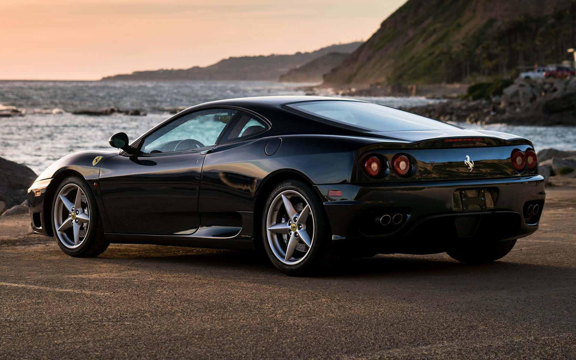 Ferrari 360 Modena 桌布 壁紙 — 一架時尚黑色法拉利停泊在日落時分的海岸公路上，背景是海洋和懸崖。