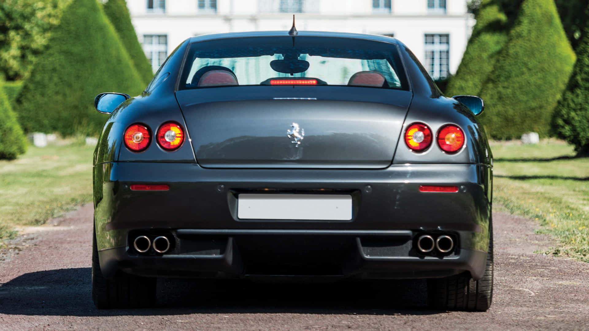 Ferrari 612 Scaglietti 桌布 壁紙 — 一架時尚黑色法拉利停泊在碎石路上，兩旁是修剪整齊的樹籬和一座宏偉的白色大宅的後視圖。