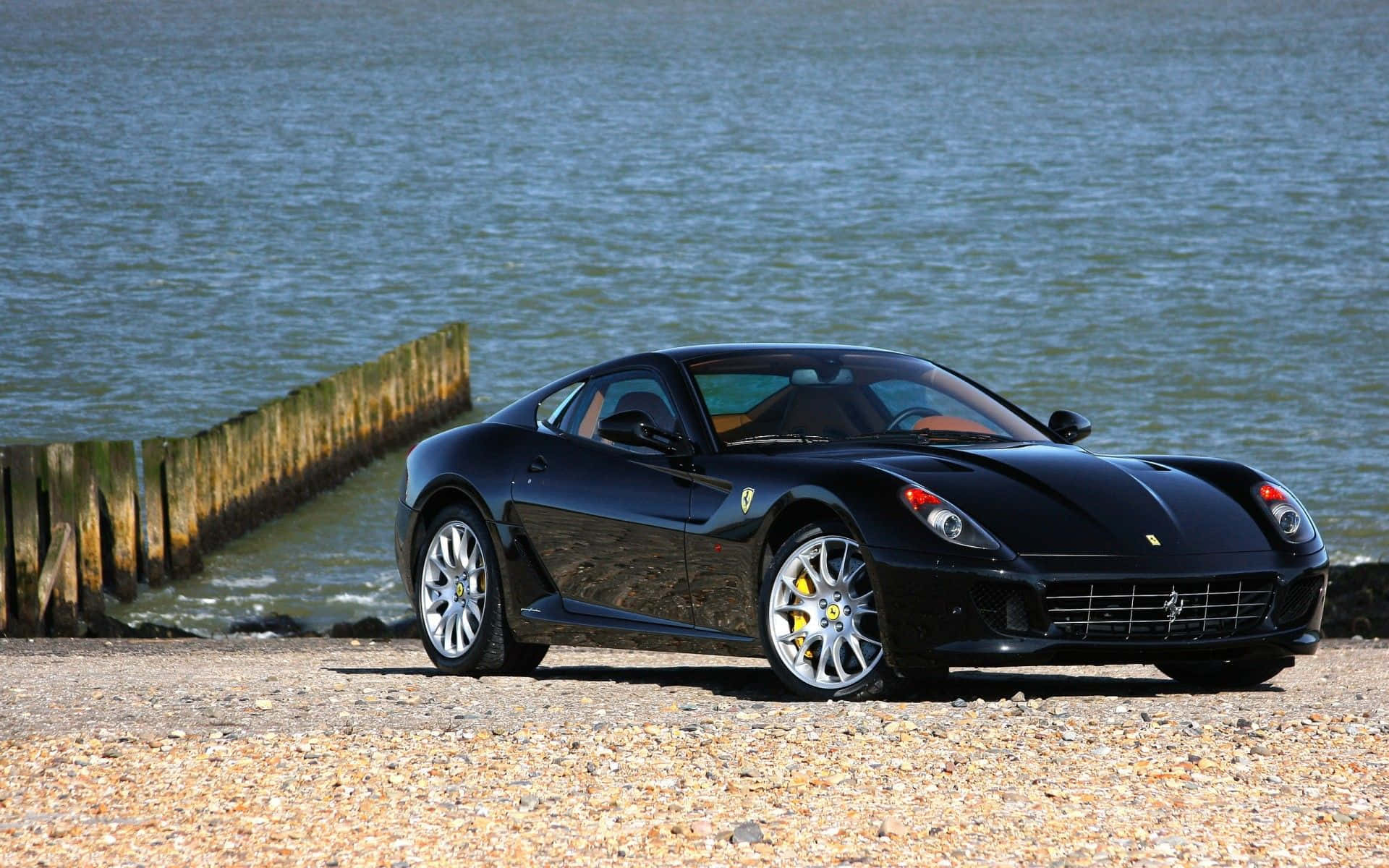 Ferrari 599 Gtb Fiorano 桌布 壁紙 — 一架時尚黑色法拉利跑車停泊在平靜水域旁邊的卵石灘上，附近有木樁。