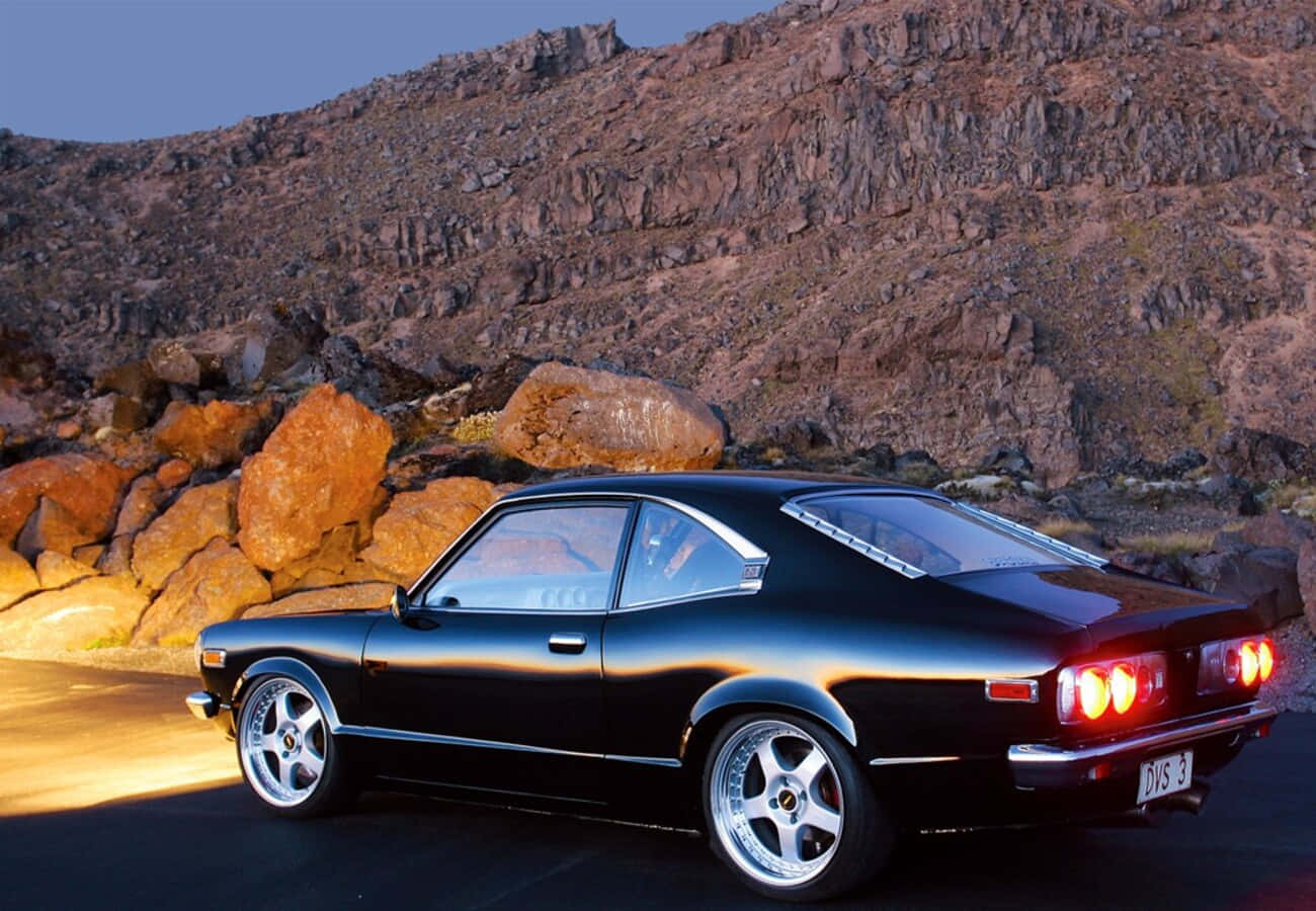 Mazda Rx-3 桌布 壁紙 — 一架時尚黑色經典跑車，帶有發光的紅色尾燈，停泊在黃昏時分的沙漠道路上，背景是崎嶇的岩石地形。