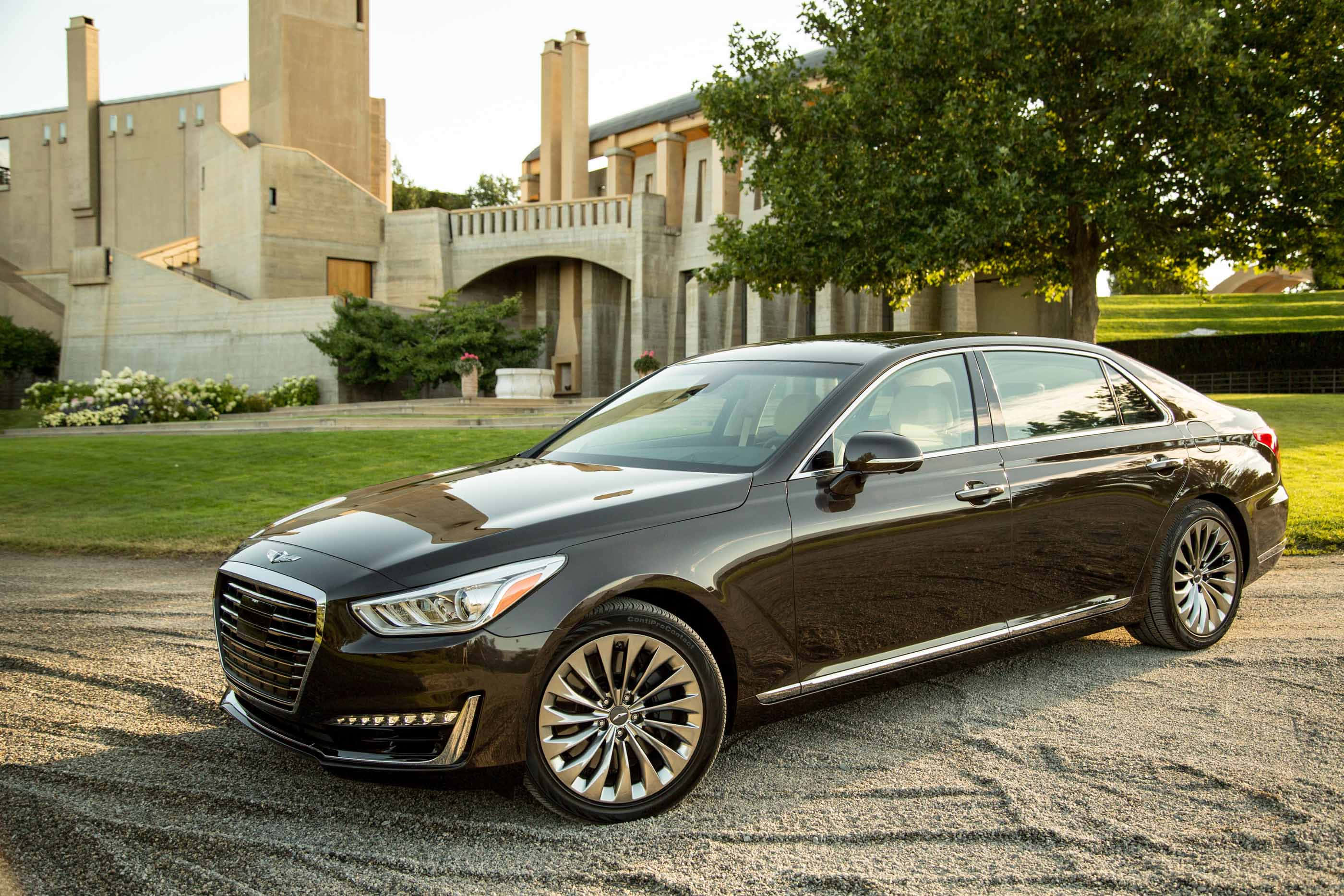 Genesis G90 桌布 壁紙 — 一架時尚黑色豪華轎車停泊在碎石路上，前方係一座現代石建築，周圍有茂密嘅綠樹同修剪整齊嘅草坪。