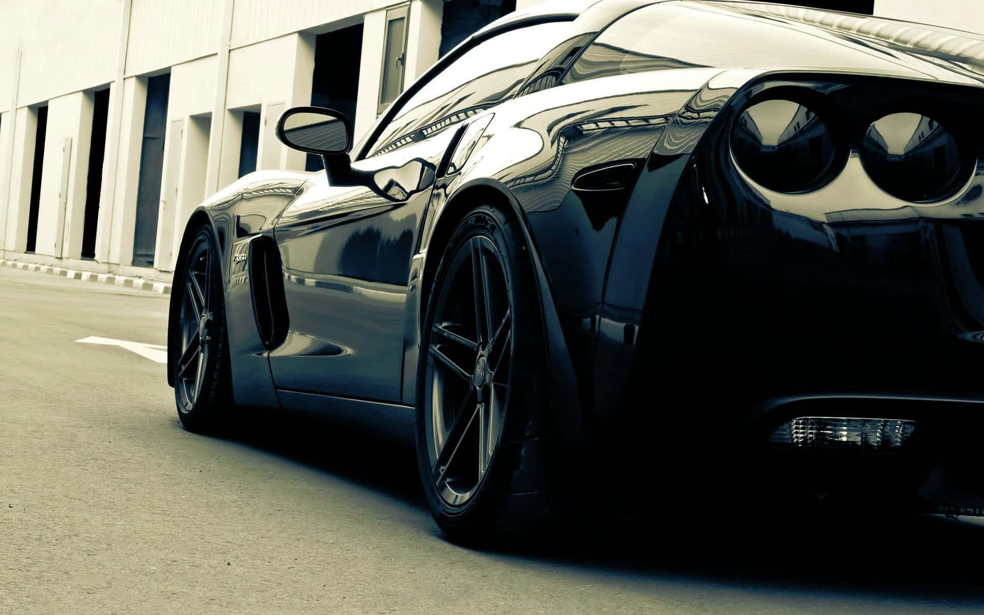 Chevrolet Corvette Z06 桌布 壁紙 — 一架時尚黑色跑車在強光下閃耀，拋光表面反射光線，停泊在現代建築旁。