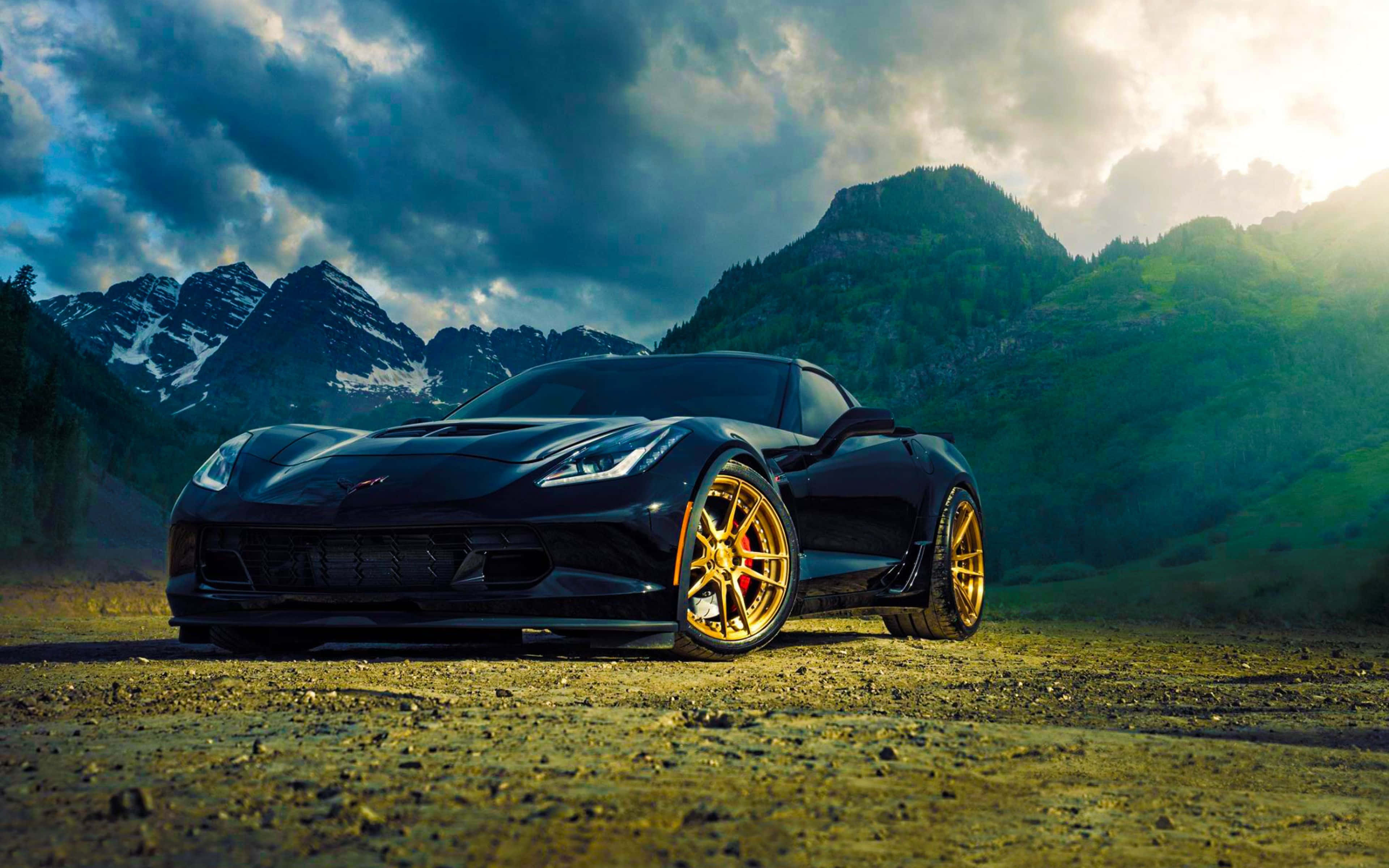 Chevrolet Corvette Z06 桌布 壁紙 — 一架時尚黑色跑車配金色輪圈，停泊在碎石路上，背景係白雪覆蓋嘅山脈同戲劇性嘅雲層。
