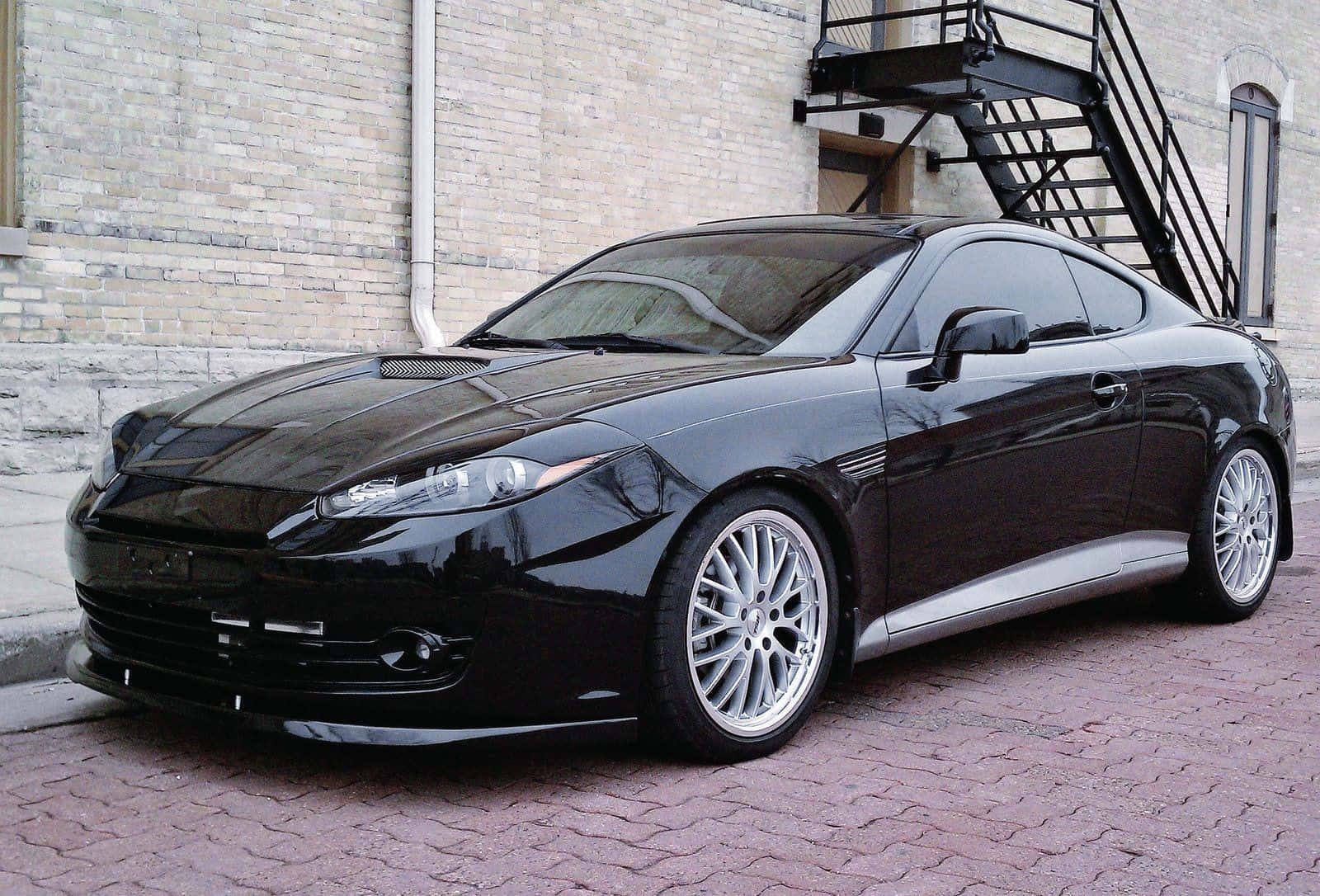 Hyundai Tiburon 桌布 壁紙 — 一架時尚黑色跑車配銀色輪圈，停泊在紅磚行人路上，旁邊係米色磚建築物，有黑色金屬樓梯。