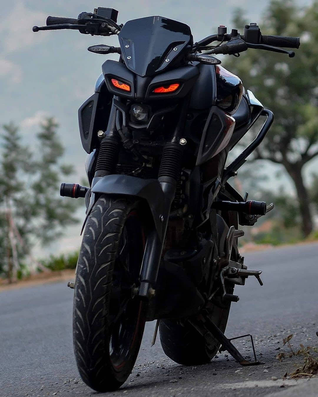 yamaha mt 15 桌布 壁紙 — 一架時尚黑色運動電單車，帶有發光的橙色眼睛，停在樹木環繞的瀝青路上，天空多雲。