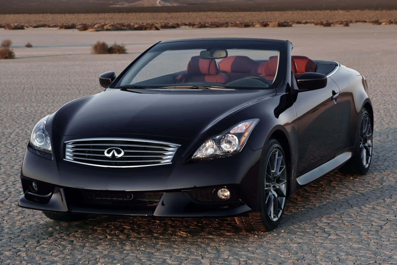 Infiniti Q60 桌布 壁紙 — 一架時尚黑色開篷跑車配紅色內籠，停泊在溫暖金色光線下龜裂的沙漠道路上。