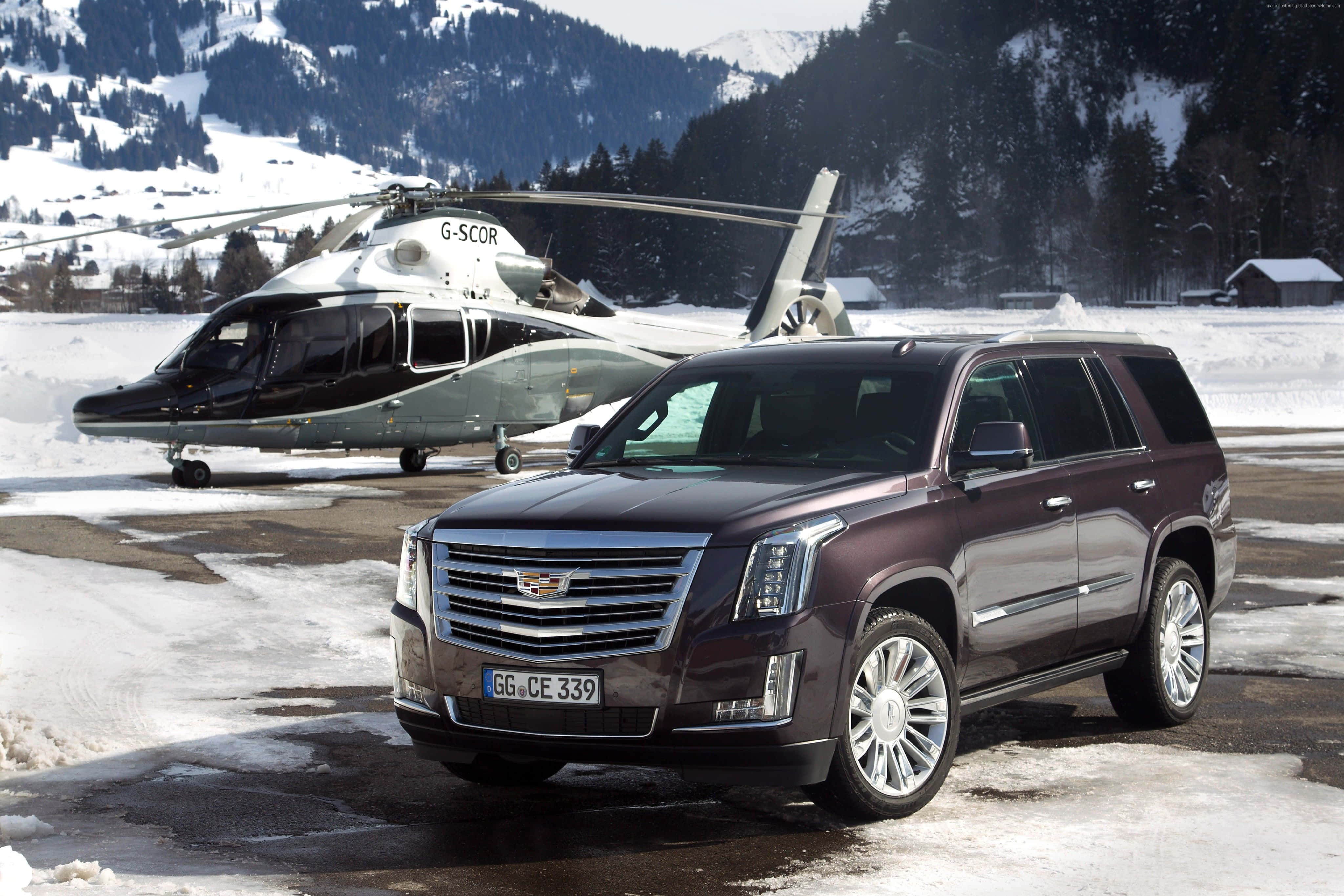 Cadillac Escalade 桌布 壁紙 — 一架棗紅色 Cadillac Escalade SUV 停泊在積雪的停機坪上，旁邊有一架黑白相間的直升機，背景是白雪覆蓋的山脈。