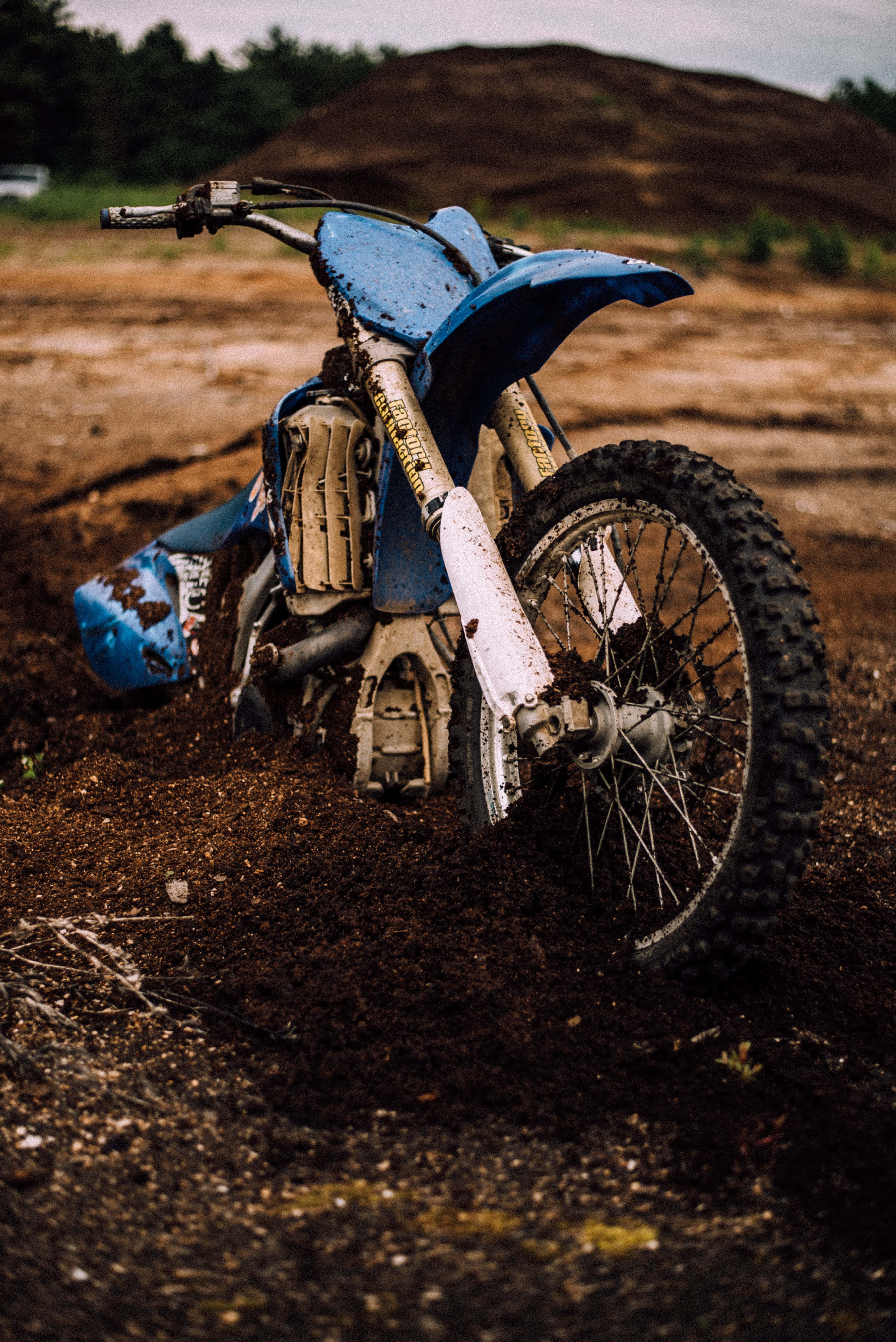 Dirtbike 桌布 壁紙 — 一架沾滿泥巴的藍色越野電單車停放在泥地上，背景有山丘。