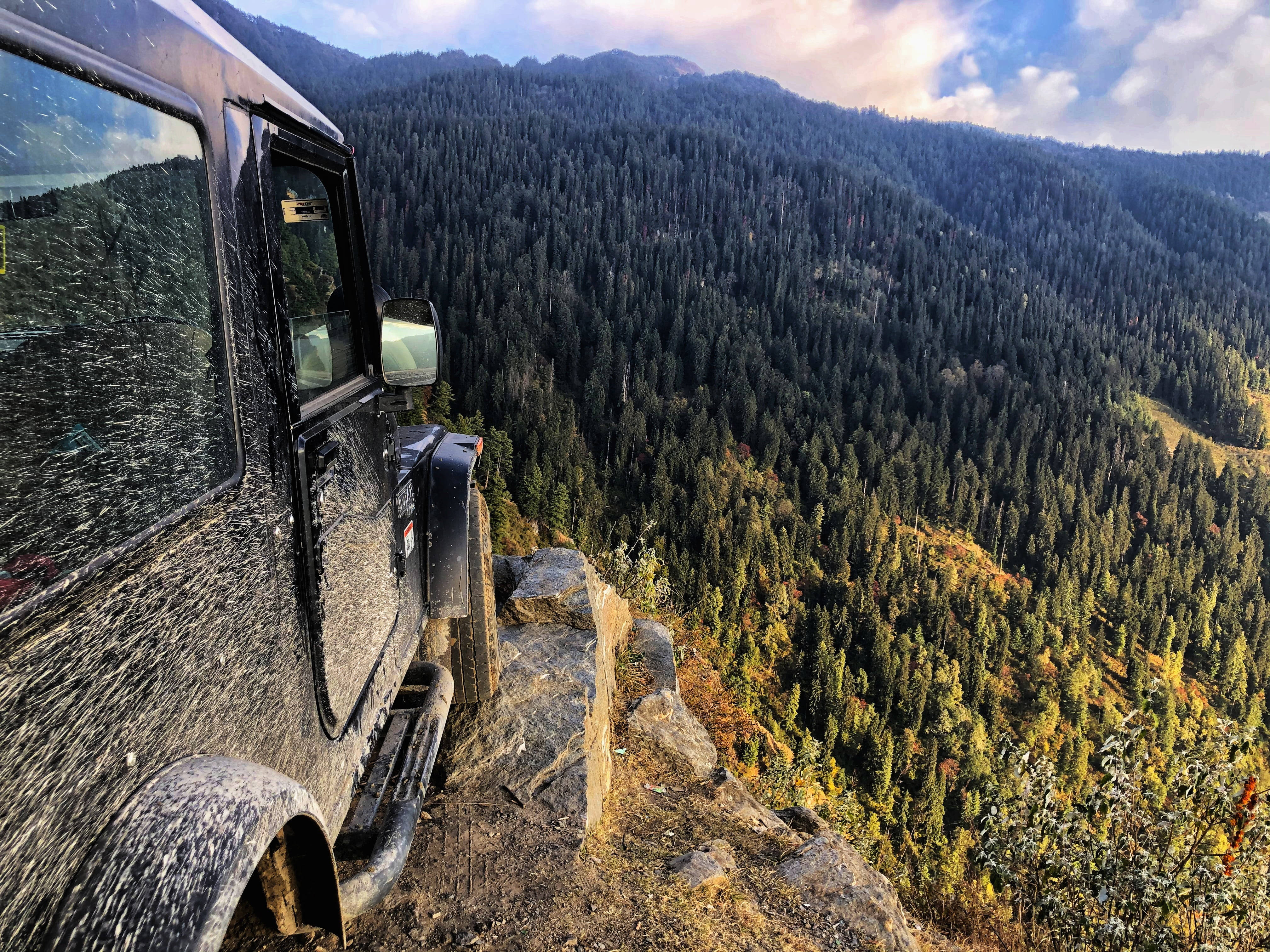Mountain 桌布 壁紙 — 一架泥濘的越野車棲息在懸崖邊緣，俯瞰著廣闊的森林山谷，沐浴在金色的日落光芒中。