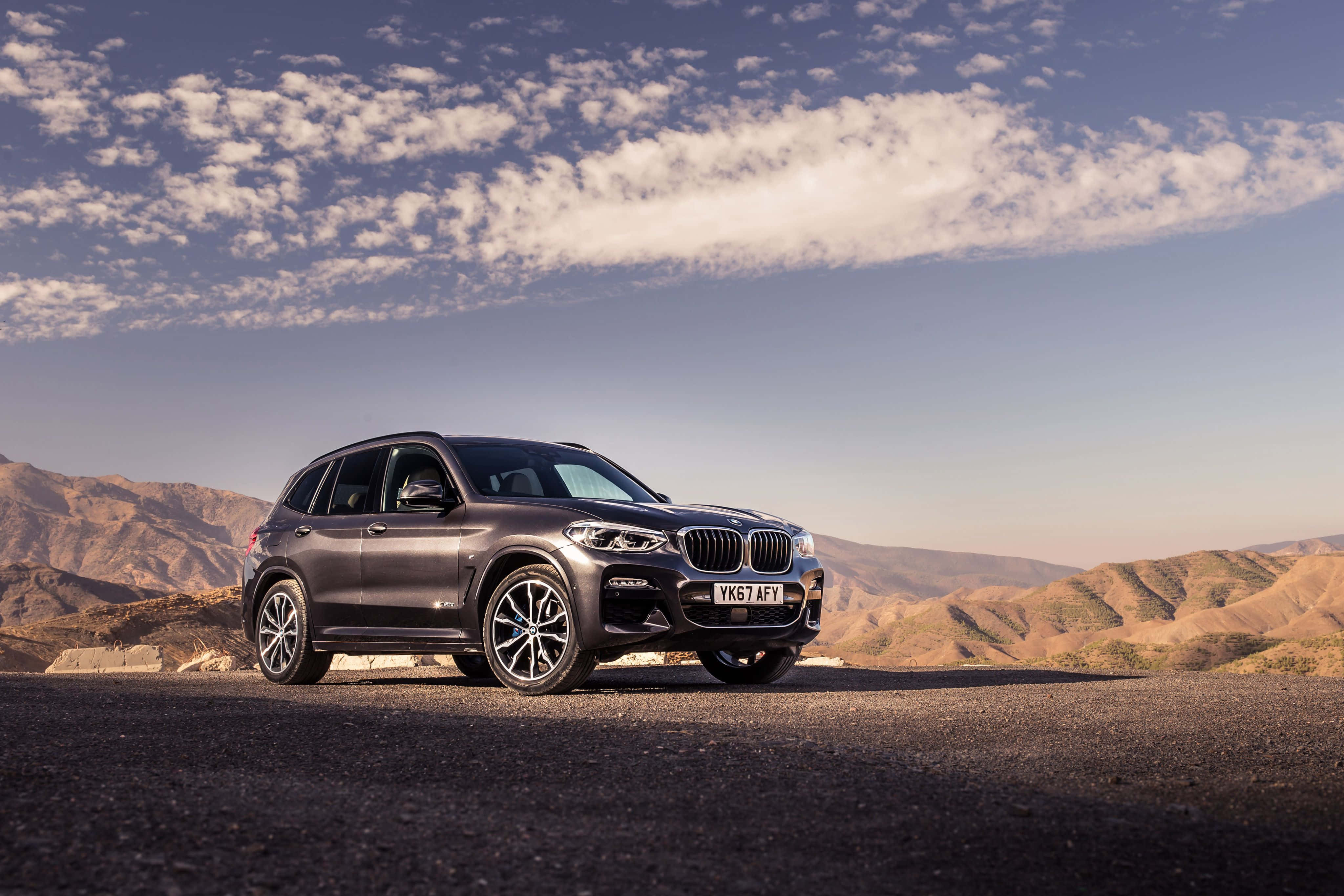 Bmw X3 桌布 壁紙 — 一架流線型啞光灰色BMW X5 SUV停泊在沙漠公路上，背景是起伏的棕色山丘和黃金時段部分多雲的天空。