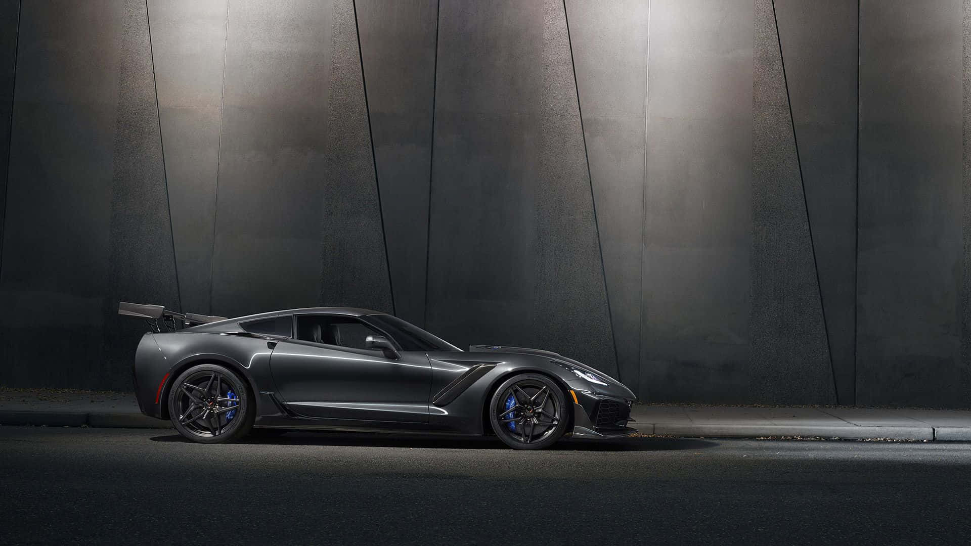 corvette 桌布 壁紙 — 一架流線型啞光灰色跑車配藍色制動卡鉗，在戲劇性聚光燈下停泊於深色工業牆前。