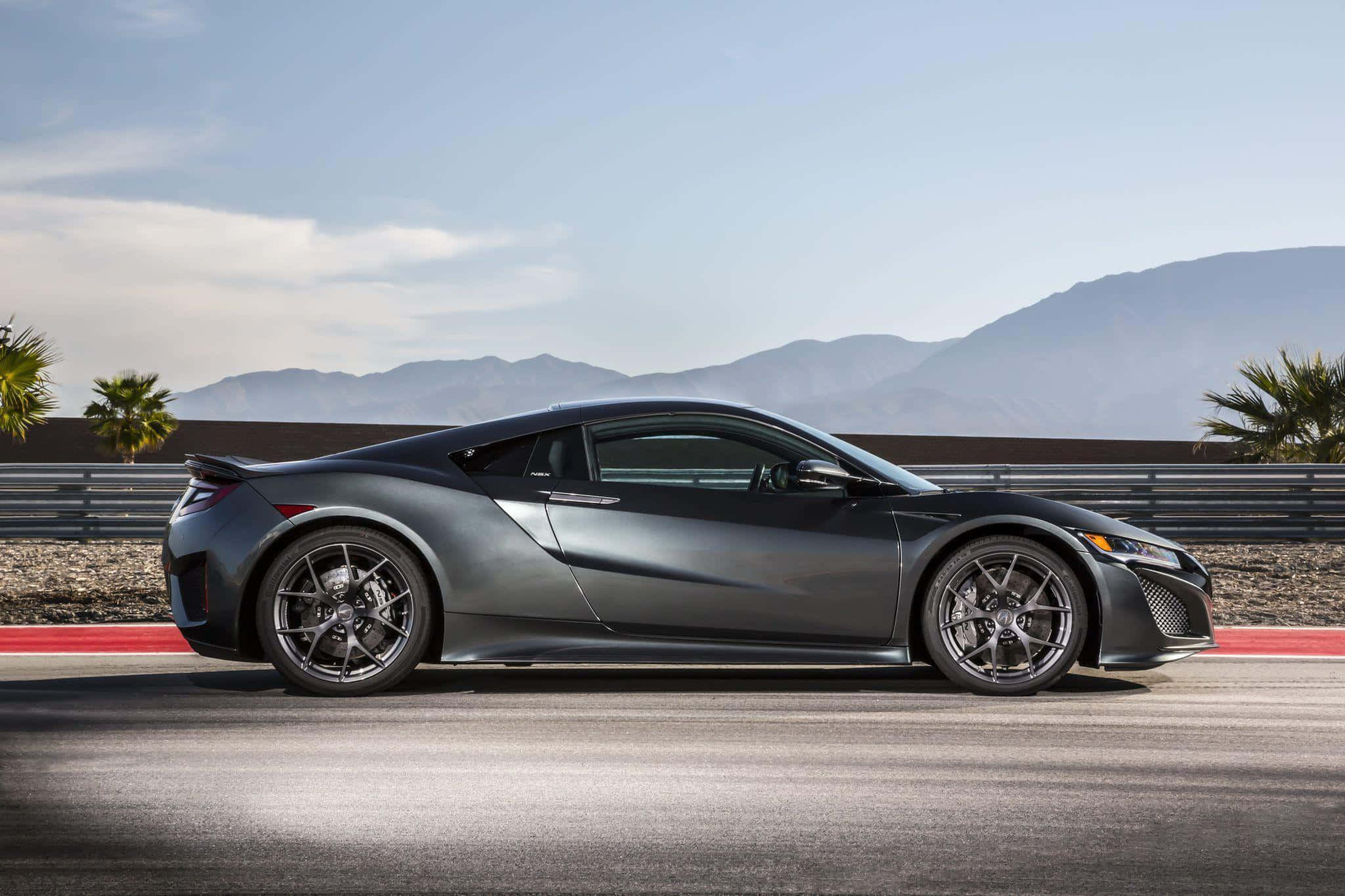 Acura Nsx 桌布 壁紙 — 一架流線型啞灰色跑車在晴朗天空下，於有遠山和棕櫚樹的沙漠賽道上行駛。