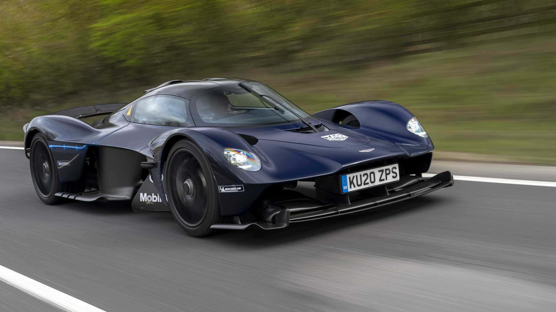 Aston Martin Valkyrie 桌布 壁紙 — 一架流線型深藍色Aston Martin Valkyrie超跑在道路上疾馳，背景是模糊的綠色樹葉。