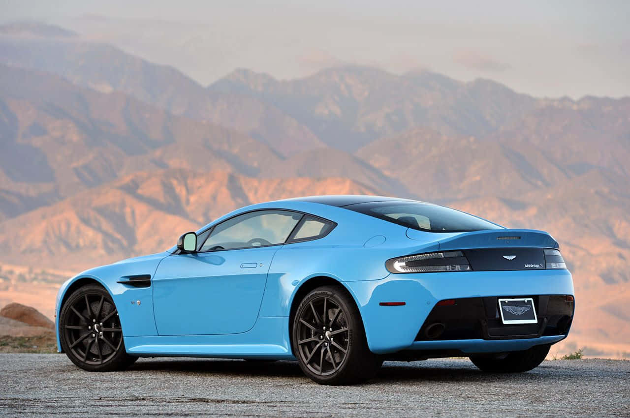 Aston Martin V12 Vantage 桌布 壁紙 — 一架流線型淺藍色Aston Martin V8 Vantage停泊在碎石觀景點，背景是朦朧的崎嶇山脈。