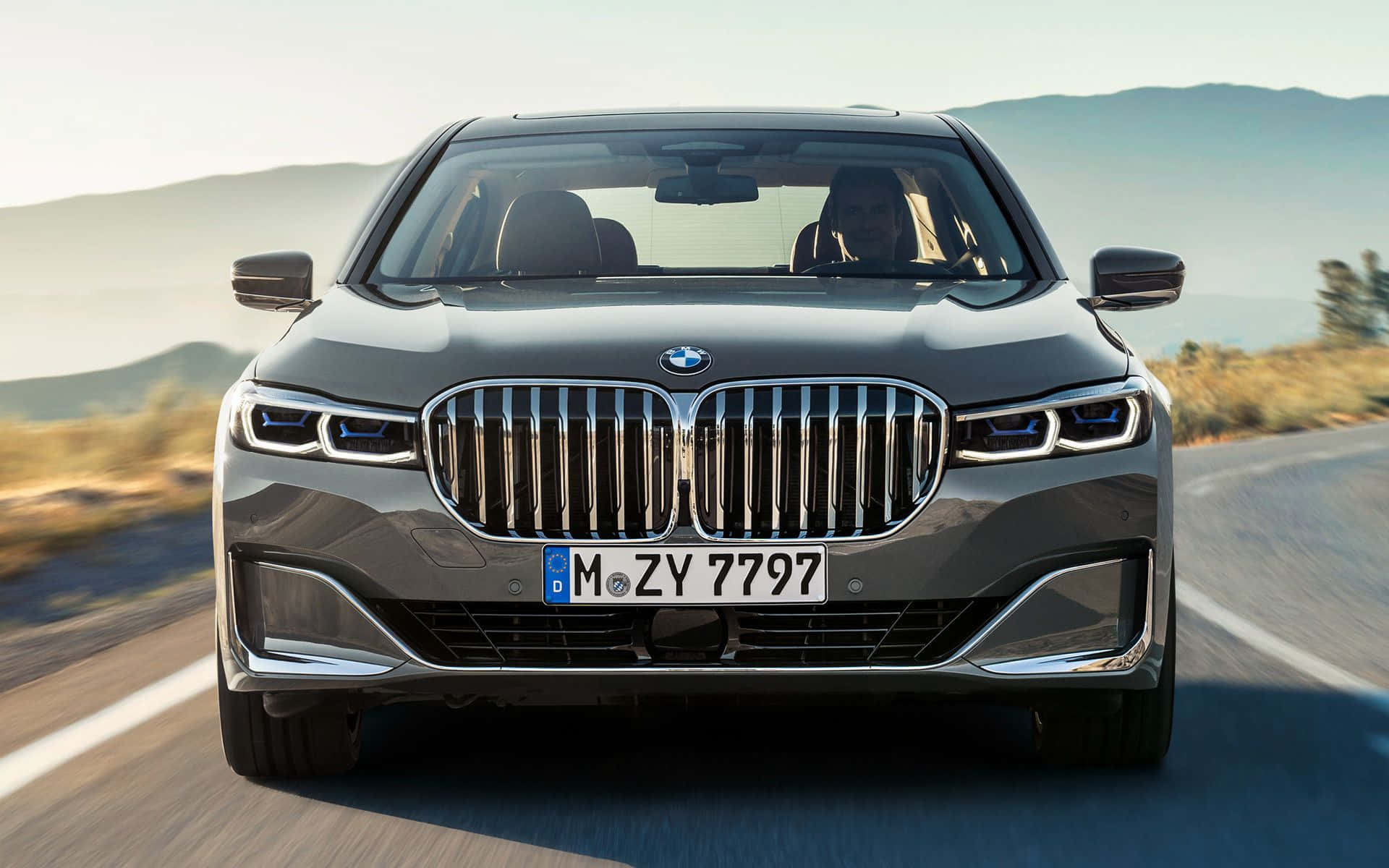 Bmw 7 Series 桌布 壁紙 — 一架流線型灰色BMW 7系房車在陽光明媚的山路上向前行駛，背景模糊。