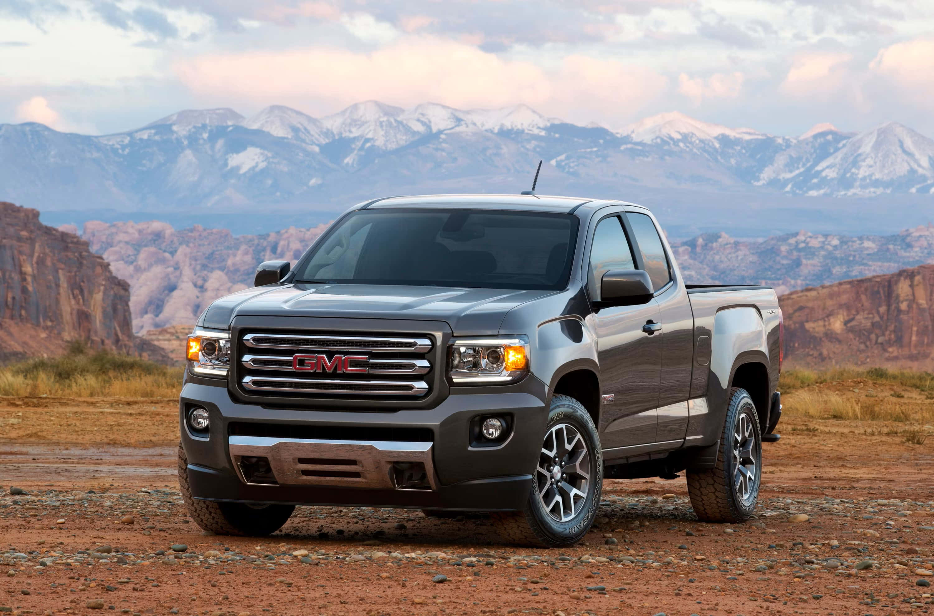 Gmc Canyon 桌布 壁紙 — 一架流線型灰色GMC Sierra皮卡車停在崎嶇的沙漠小徑上，背景中白雪覆蓋的山脈在柔和暮色下巍峨聳立。