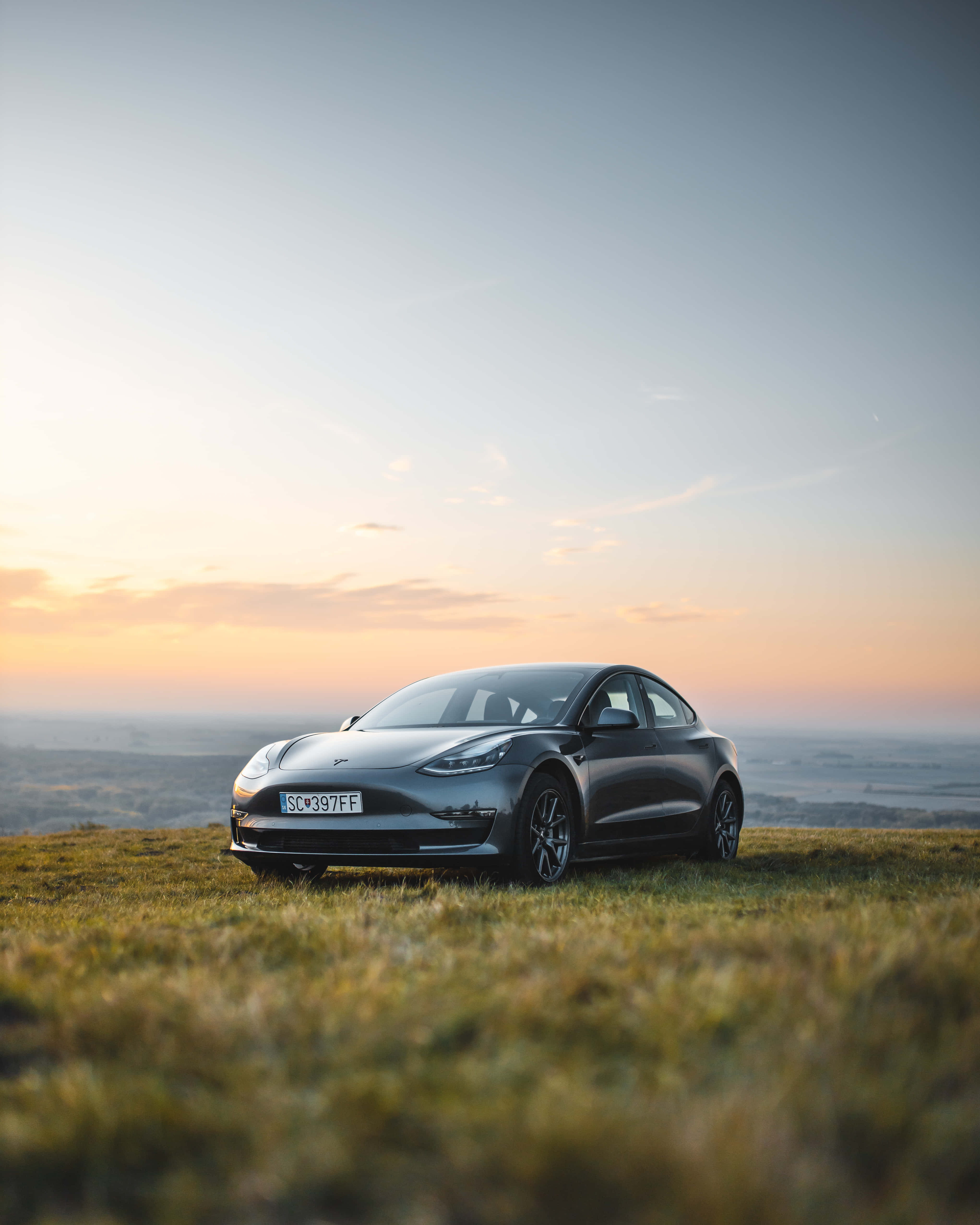 tesla 桌布 壁紙 — 一架流線型灰色Tesla Model 3停喺日落時分嘅草地上，上面係柔和嘅橙色同藍色天空。