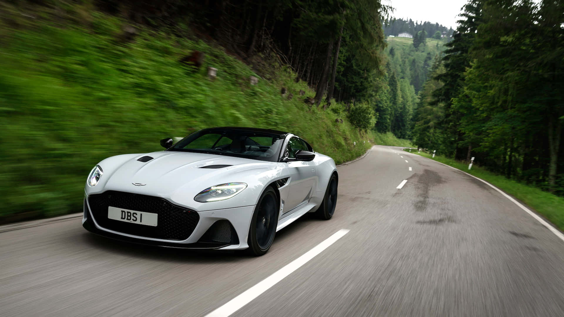 Aston Martin Dbs Superleggera 桌布 壁紙 — 一架流線型白色Aston Martin DBS Superleggera在蜿蜒的森林道路上高速行駛，動態模糊效果凸顯速度感。