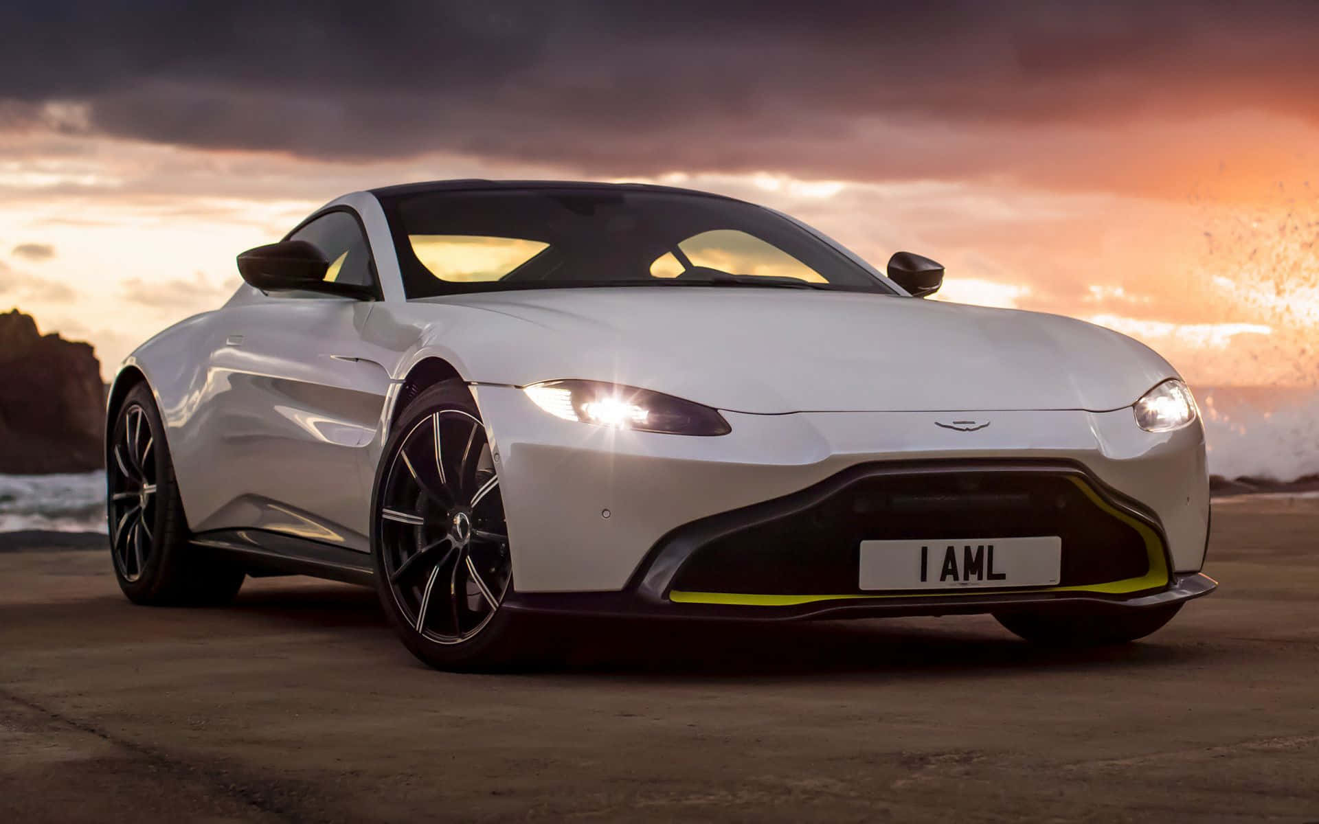 Aston Martin V12 Vantage 桌布 壁紙 — 一架流線型白色Aston Martin跑車在戲劇性的日落天空下，在拍岸的海浪旁閃閃發光。